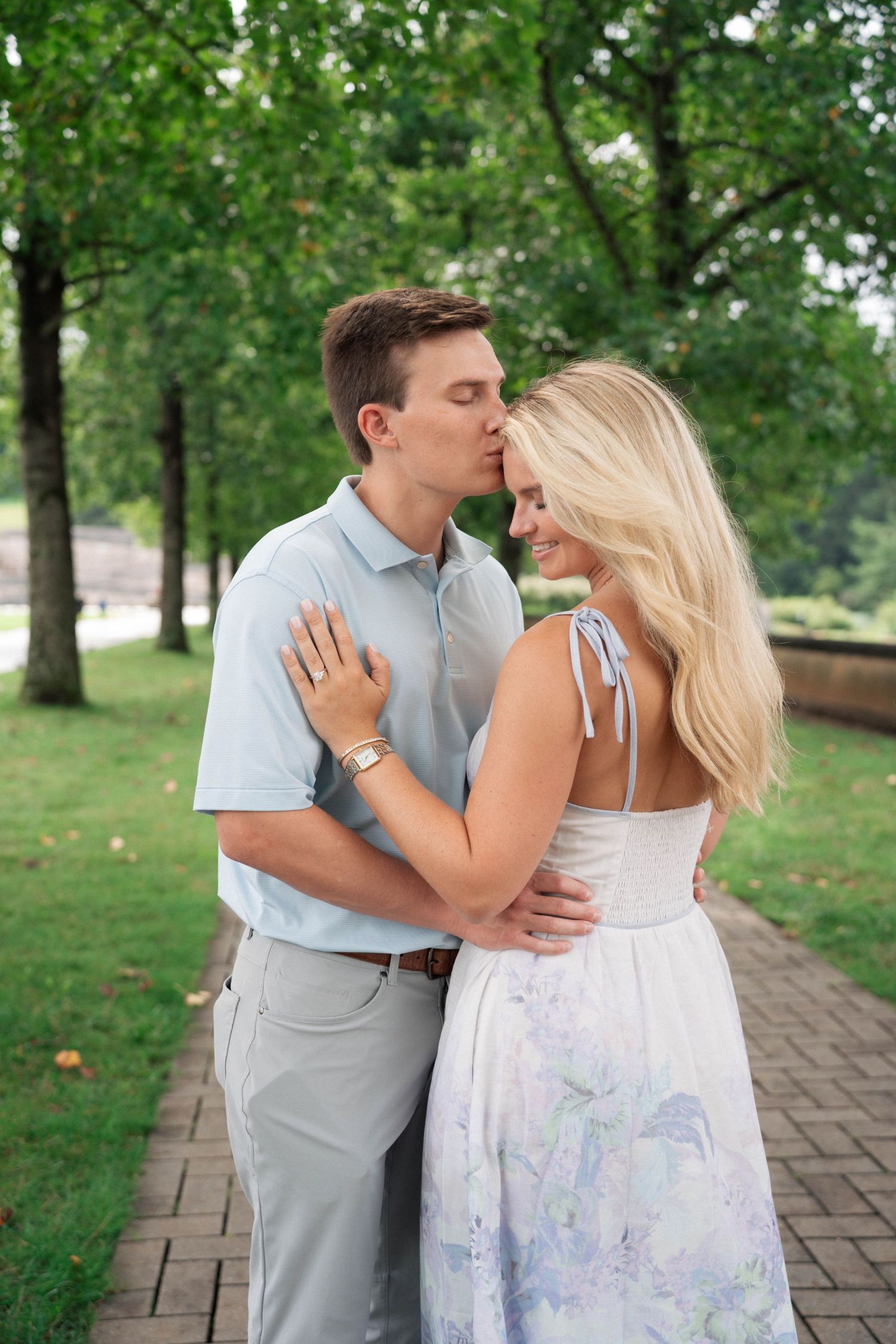 Couple sharing a tender kiss in a lush green park, woman displaying engagement ring in floral dress.