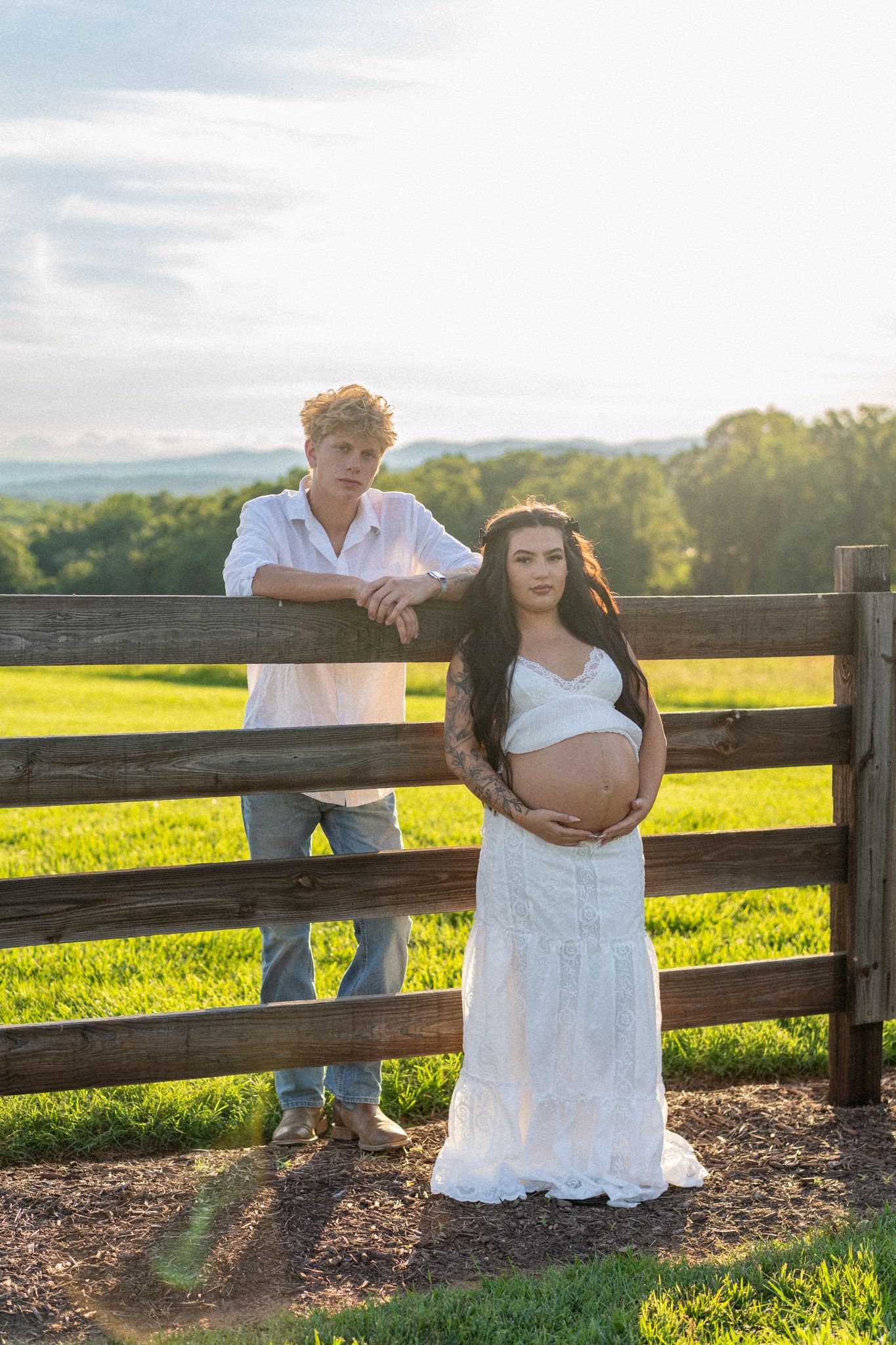 Pregnant woman in white dress cradles baby bump beside wooden fence at golden hour with partner.