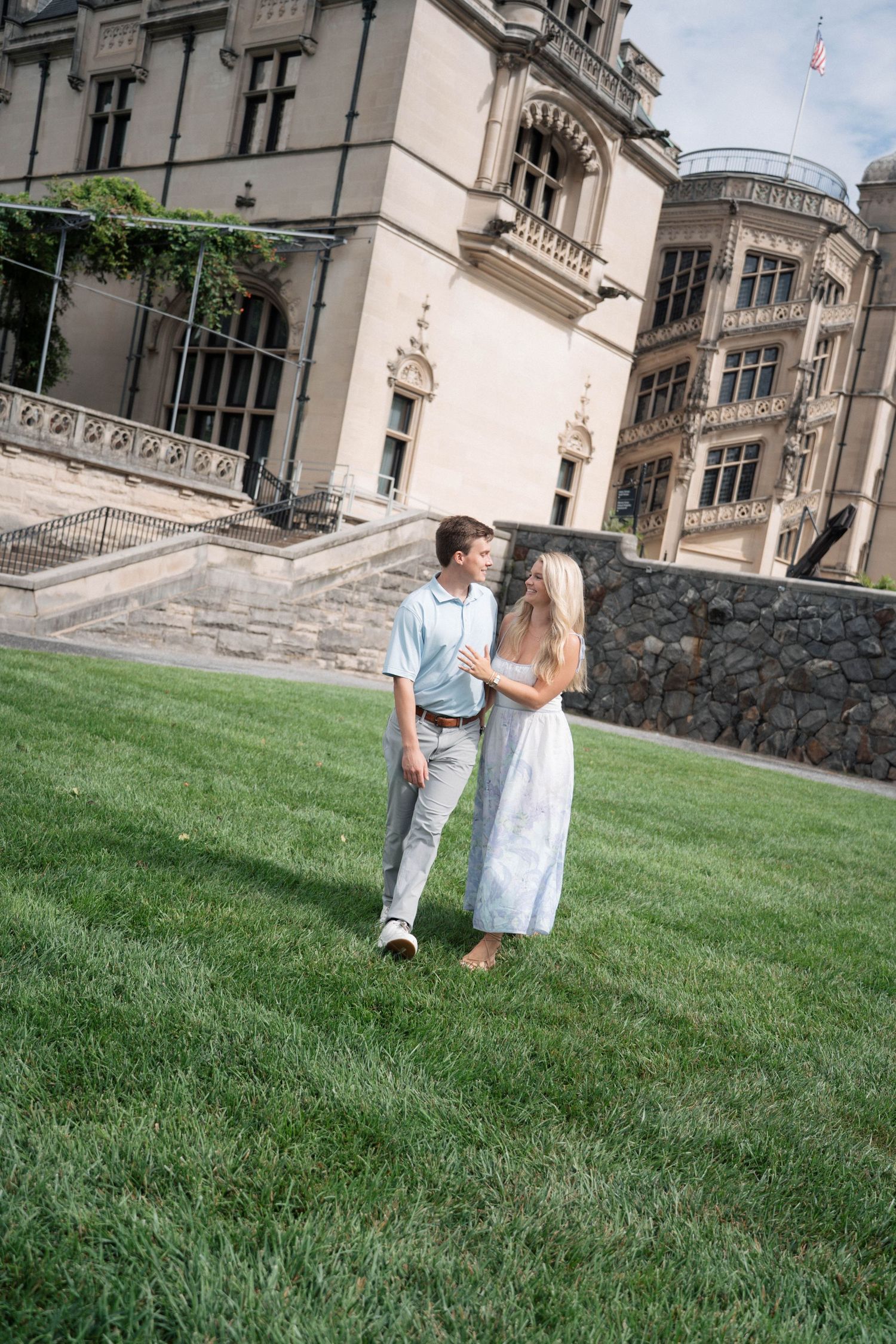Couple holding hands and smiling at each other on green lawn in front of elegant historic mansion.