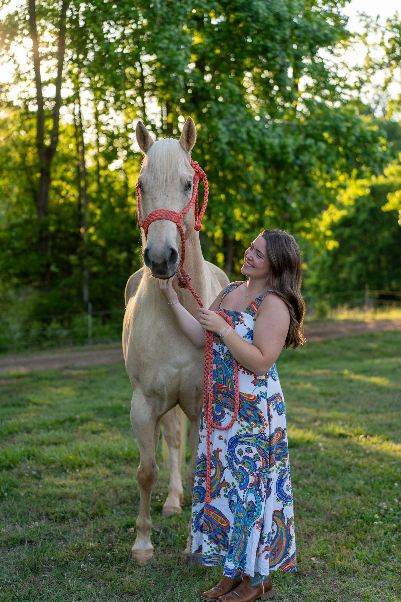 A smiling woman in a floral dress stands beside a palomino horse with a red halter in a sunny green field.