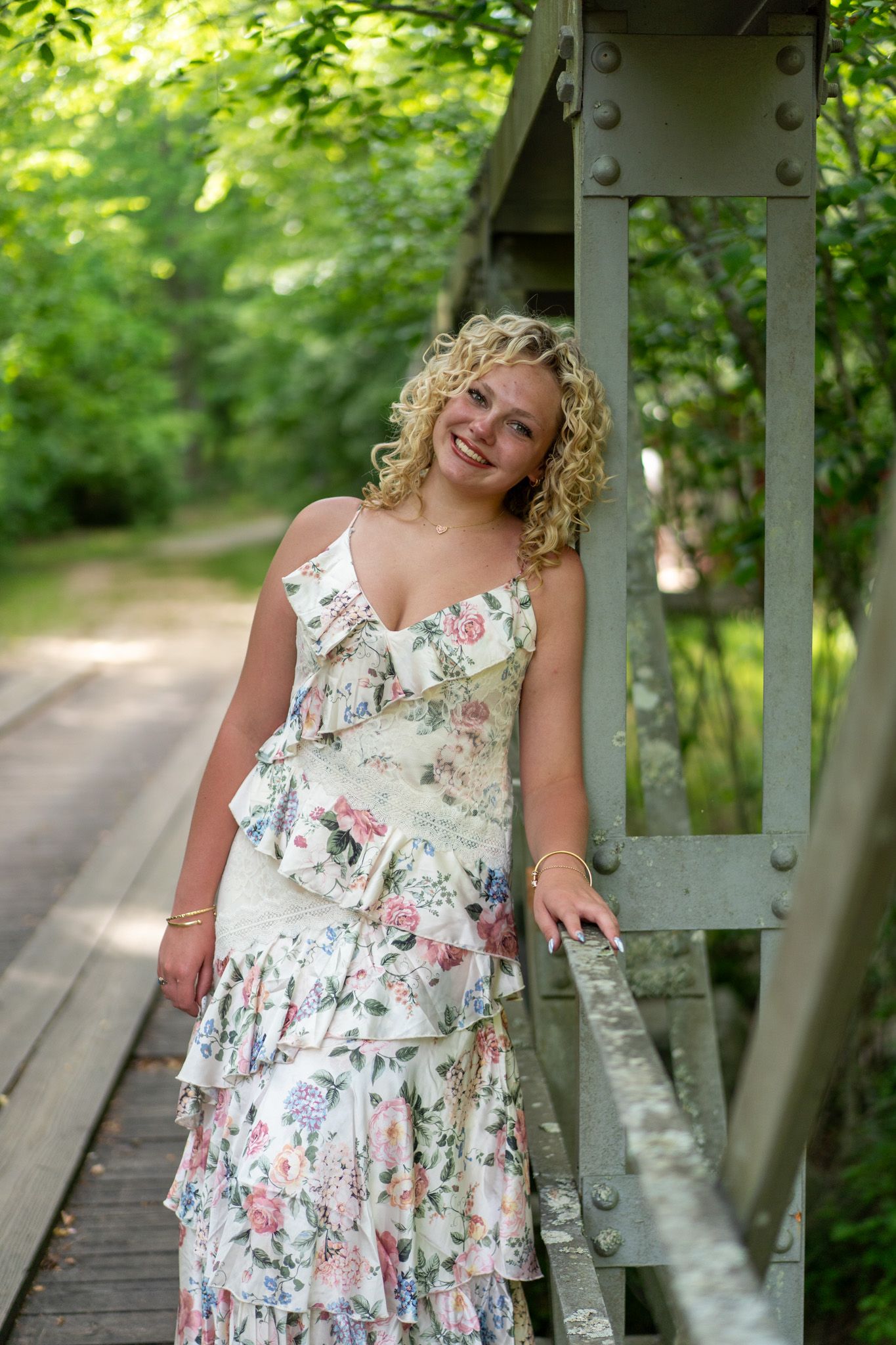 Smiling blonde curly-haired woman in floral dress posing on a bridge surrounded by lush green trees.