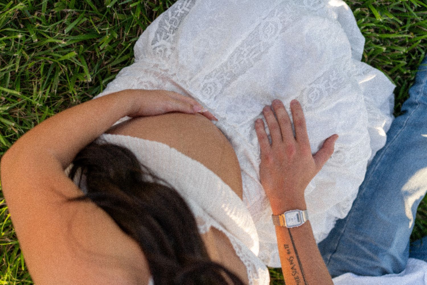 Aerial view of pregnant woman in white lace dress lying on green grass with partner's hand on her back.