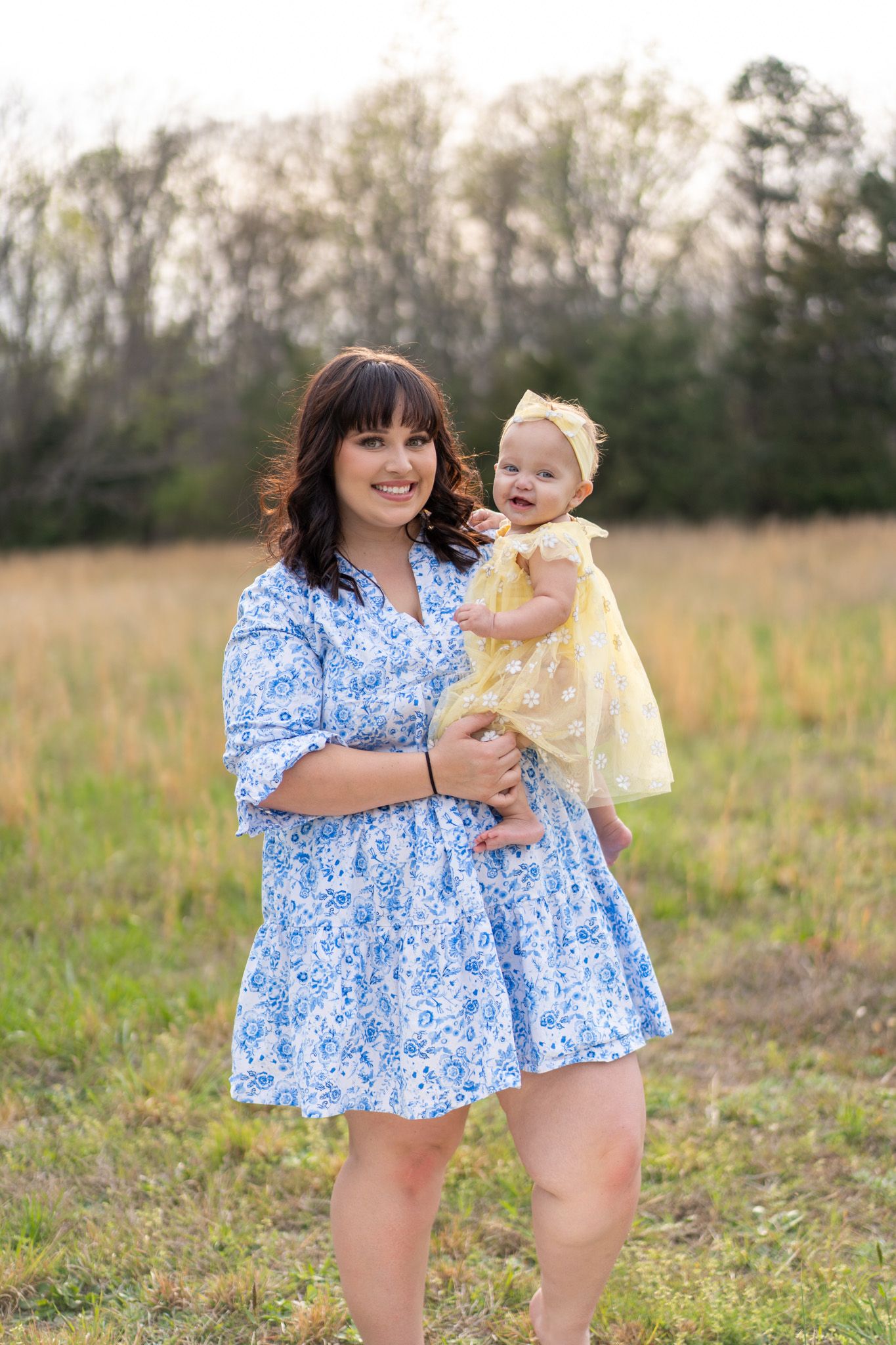 Smiling mother in blue floral dress holding her baby girl in a yellow dress outdoors.
