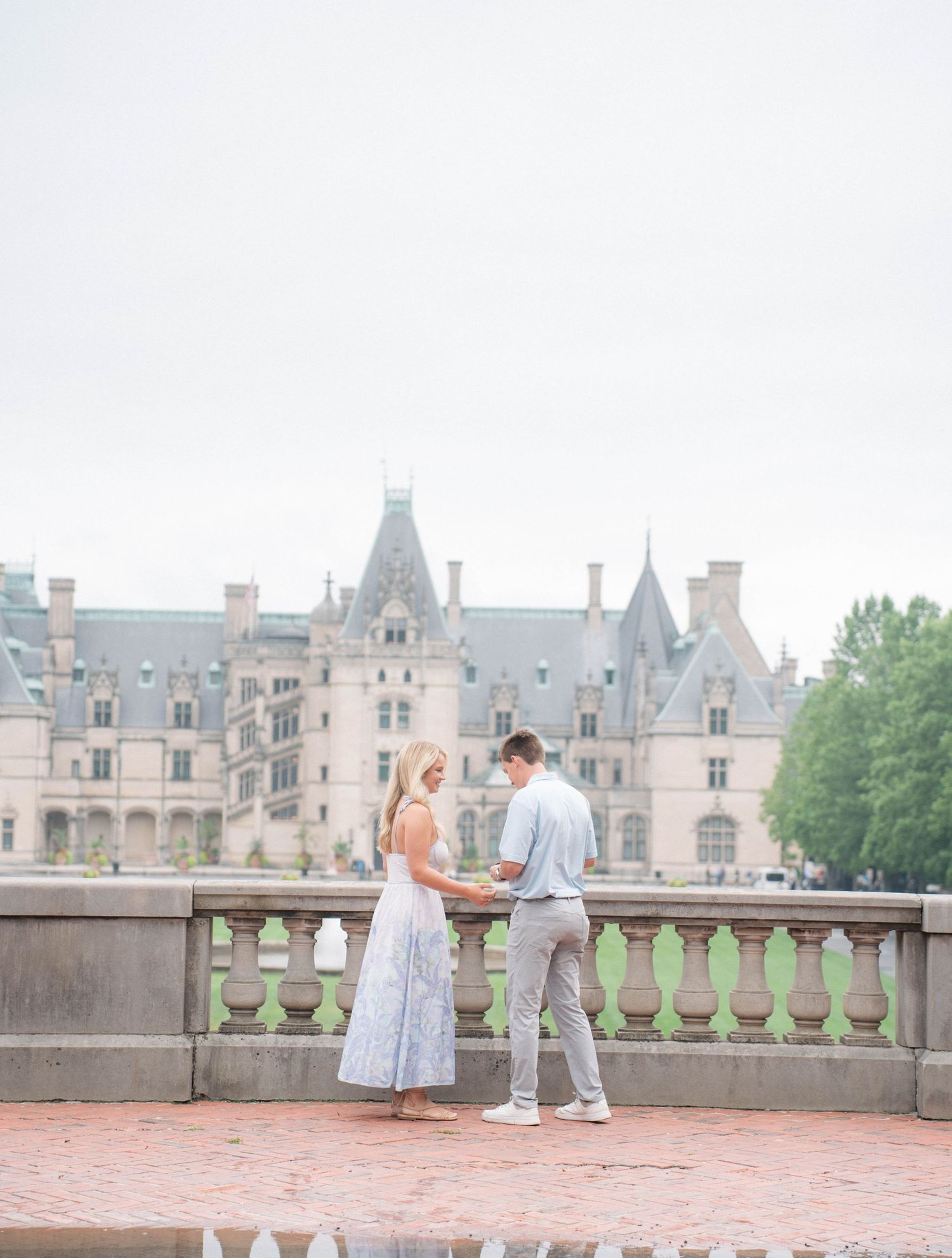 Couple holding hands on ornate stone terrace with grand Biltmore Estate castle in background.