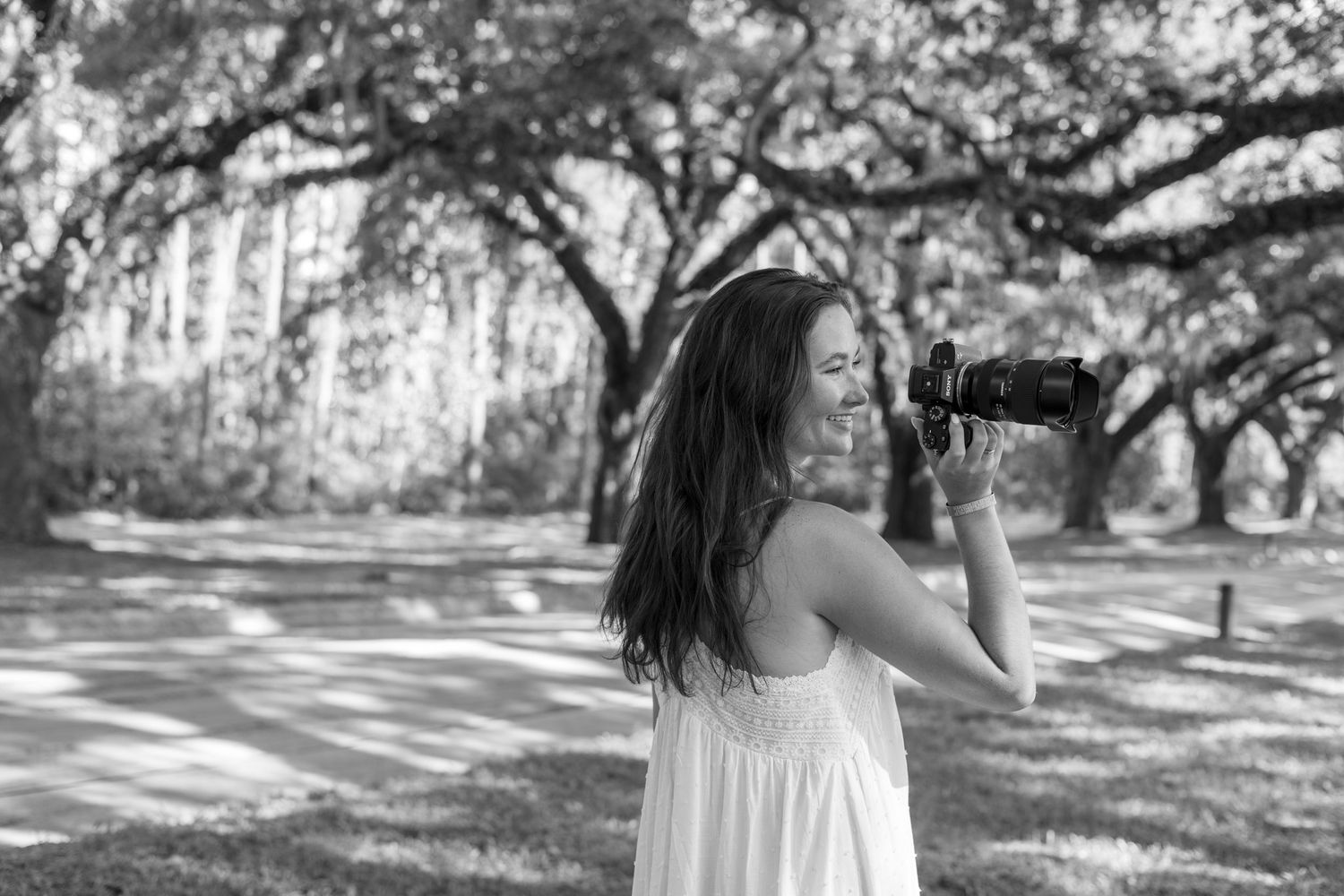 Young woman in white dress photographing nature in black and white park setting.