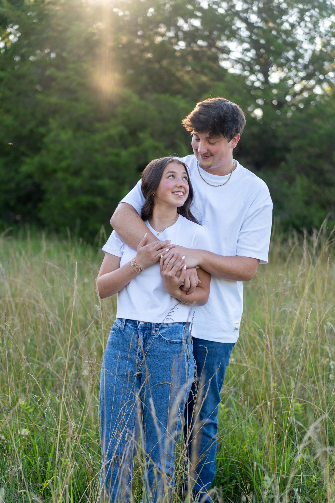 A happy young couple in white shirts and jeans embraces in a golden sunlit meadow with tall grass.