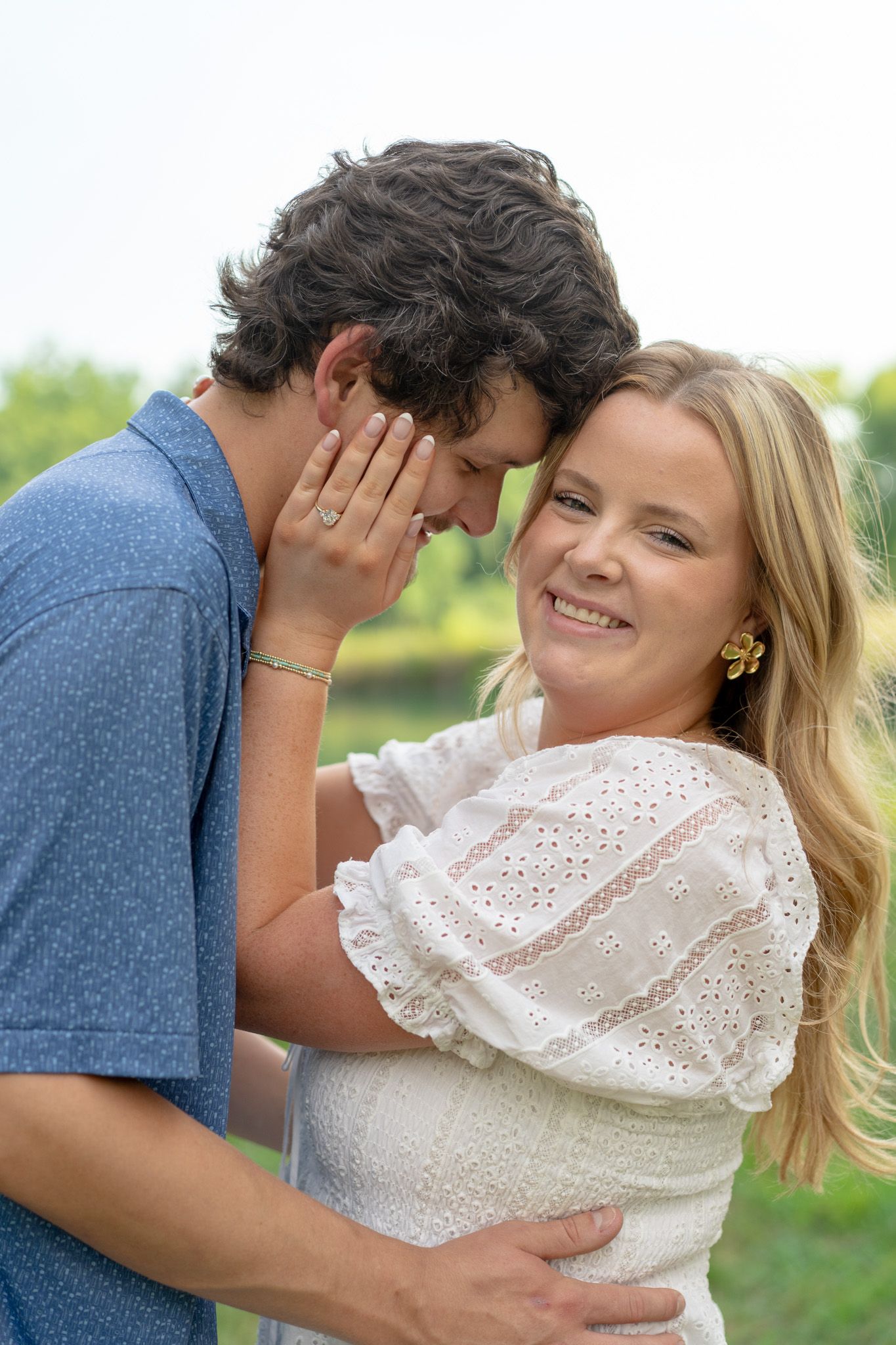 Smiling blonde woman in white dress embraces dark-haired man in blue shirt outdoors in romantic couple portrait.
