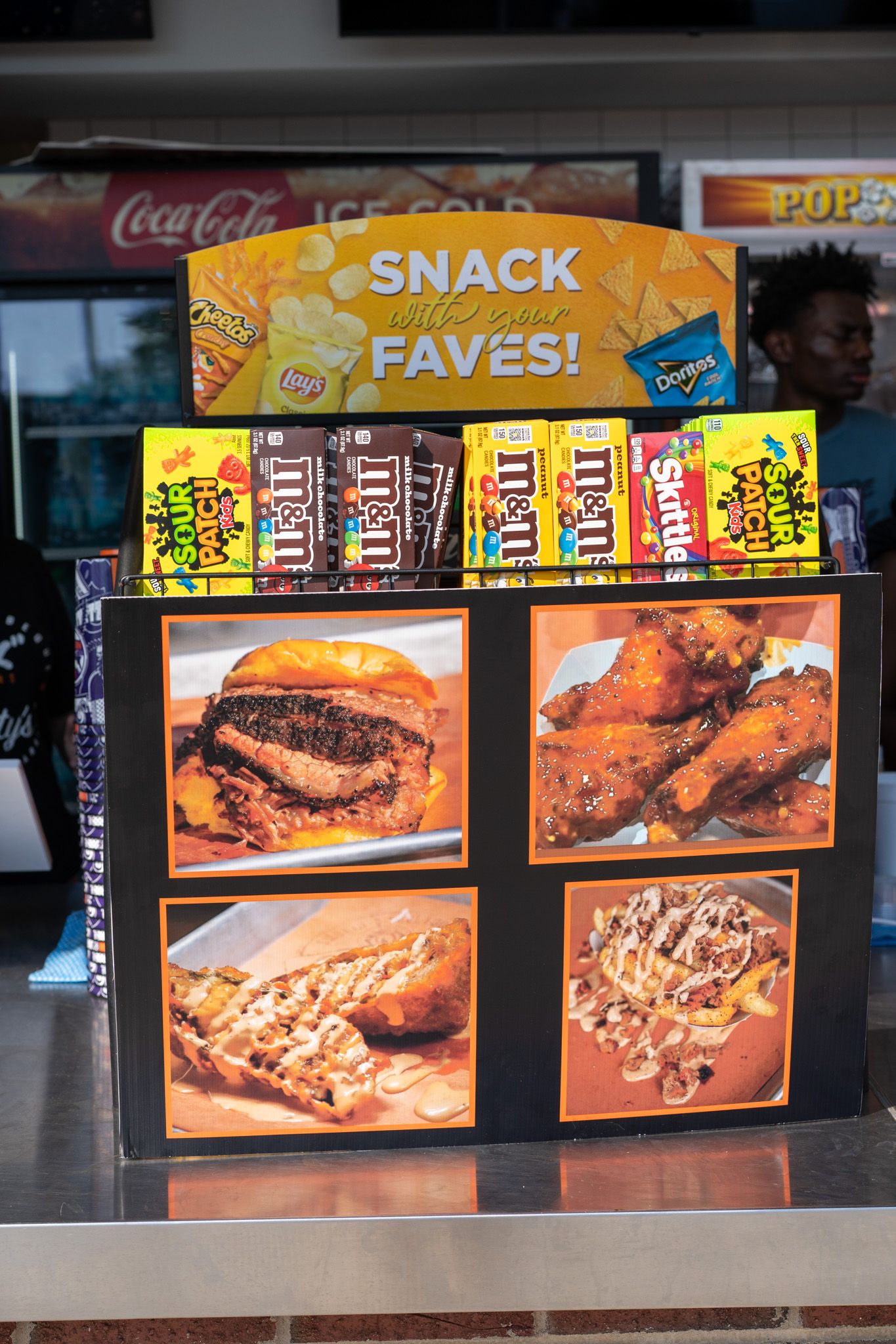 A snack display stand featuring M&Ms, Skittles, and Sour Patch Kids alongside food photos of burgers, wings, and loaded fries.