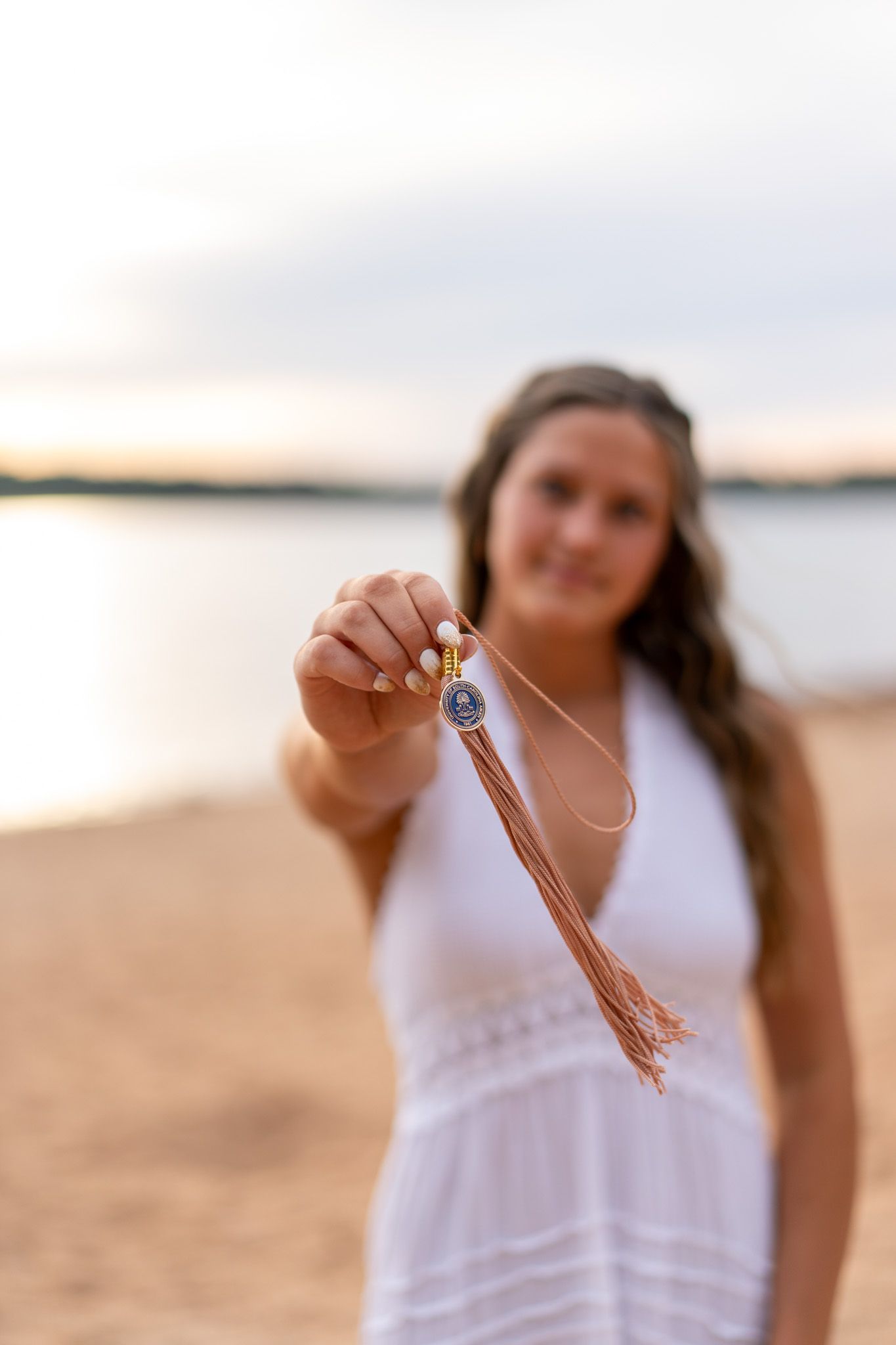 Young woman in white dress on beach holding a decorative tassel necklace pendant toward camera at sunset.