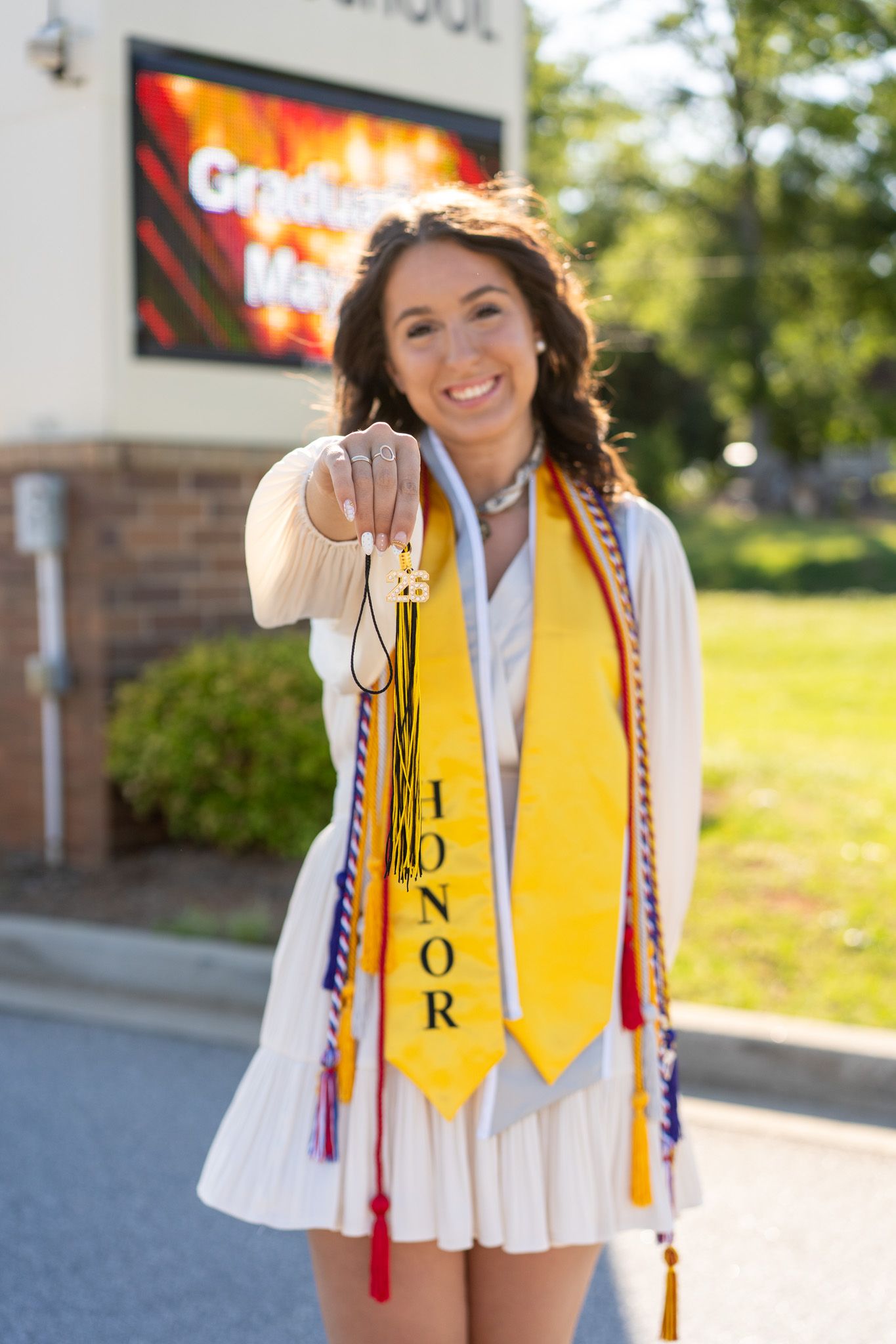 Smiling female graduate wearing honor stole and colorful cords holds up a graduation tassel outdoors.