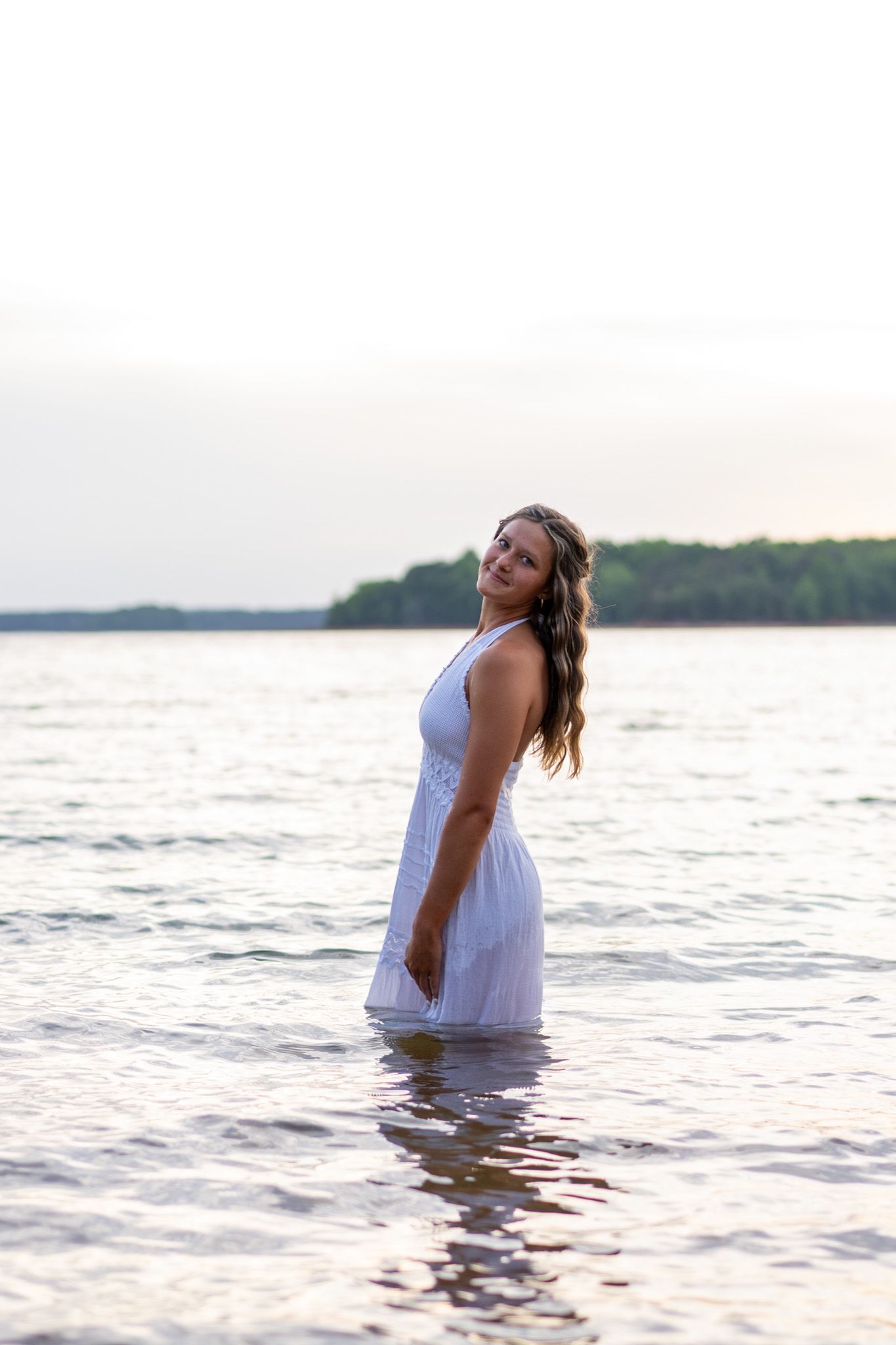 Young woman in white dress standing in calm lake water at sunset with trees in background.
