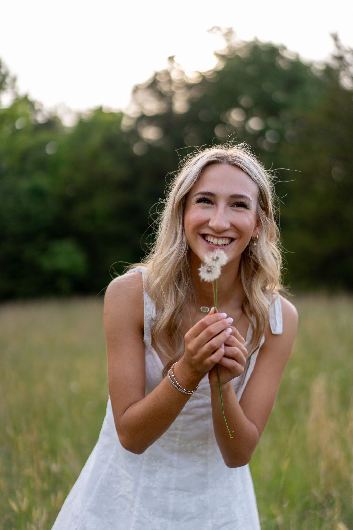 Smiling blonde woman in white dress holding a dandelion in a sunny green field.