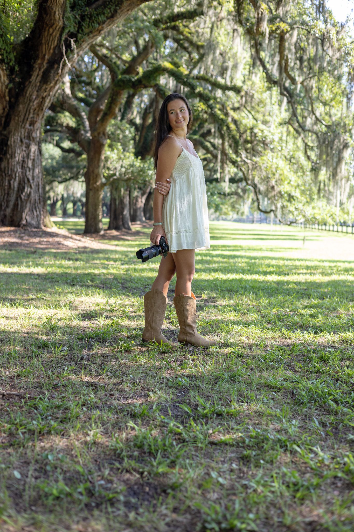 Woman in white dress and cowboy boots holding camera standing among moss-draped oak trees in a sunny park.