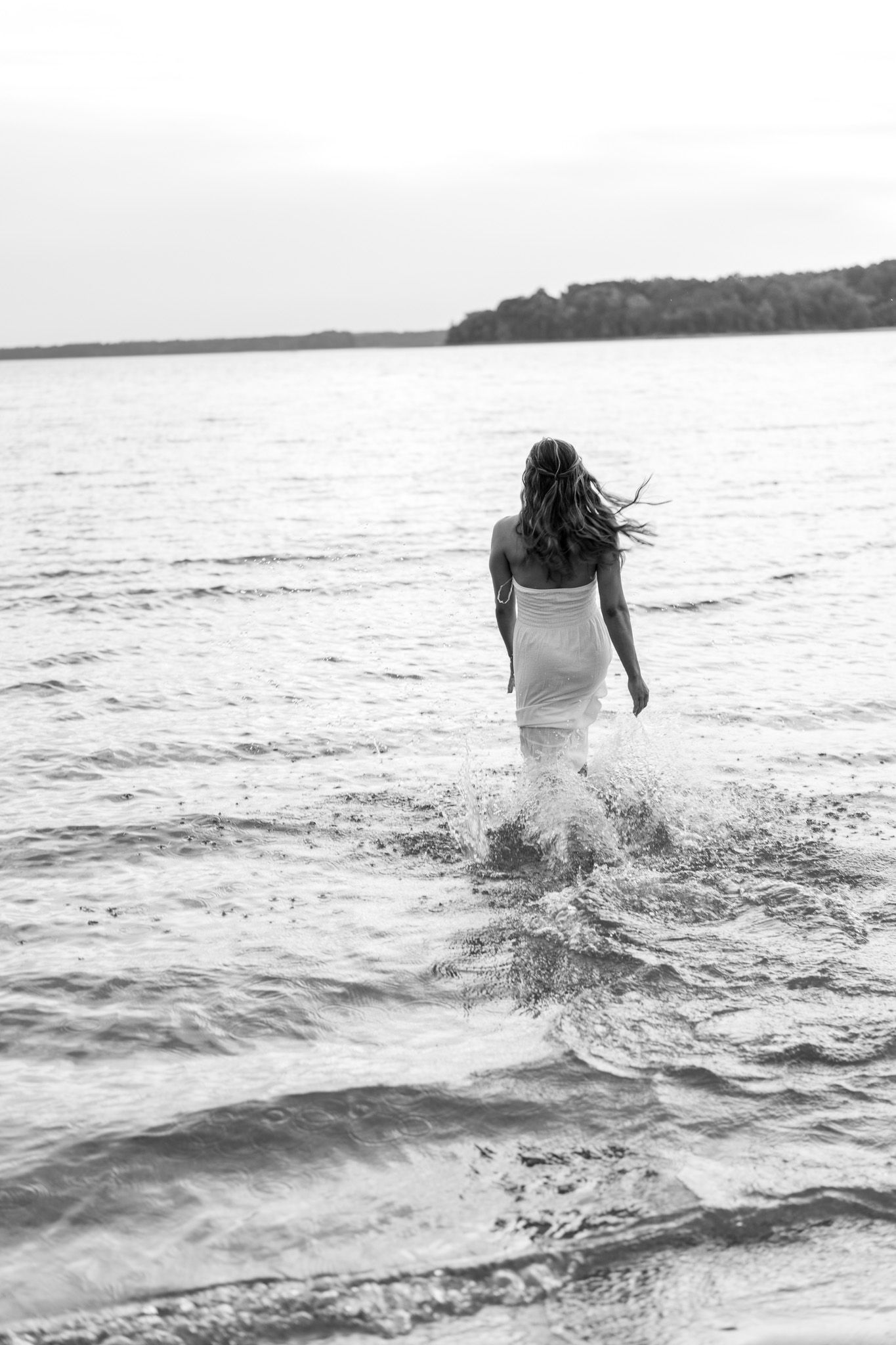 Woman in white dress wading into a calm lake, captured in a black and white photograph.