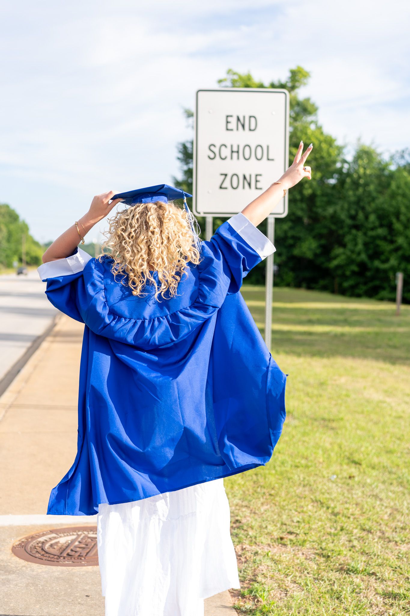 A graduate in blue cap and gown poses in front of an End School Zone sign, celebrating with arms raised.