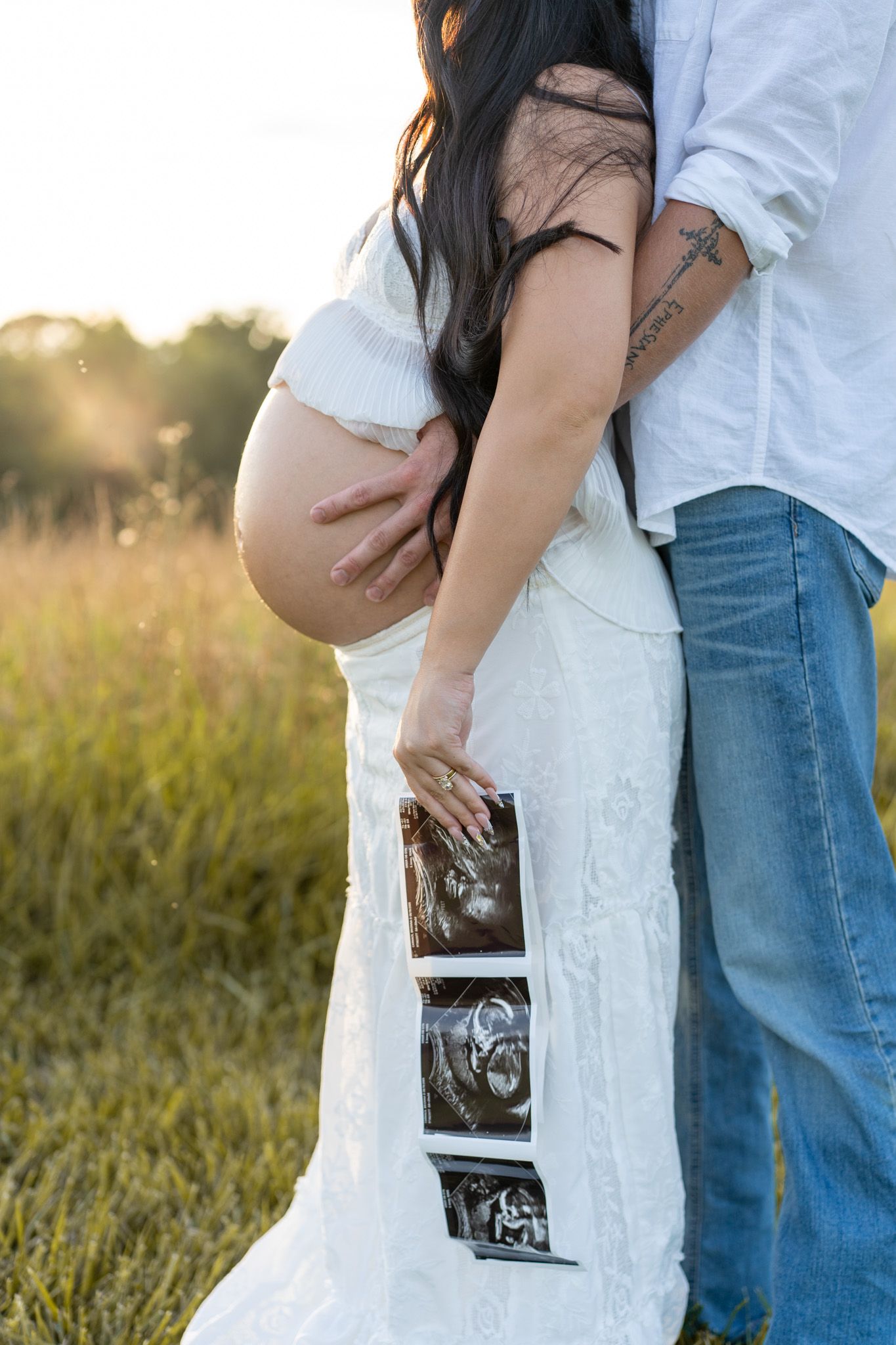 Couple embracing in golden sunset field, pregnant woman holding ultrasound photos against her baby bump.