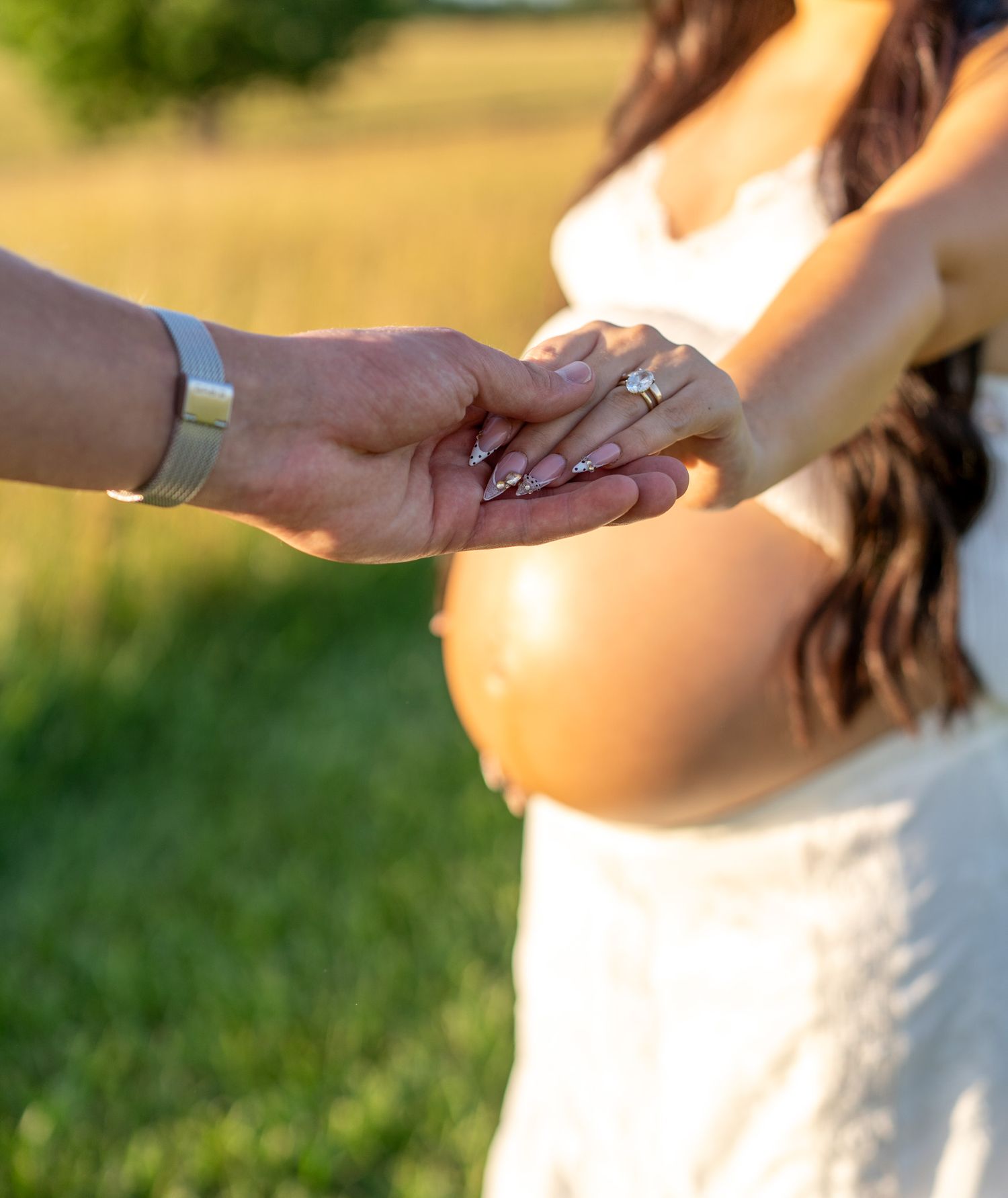 Pregnant woman in white dress holding hands with partner outdoors in golden sunlight, showing engagement ring.
