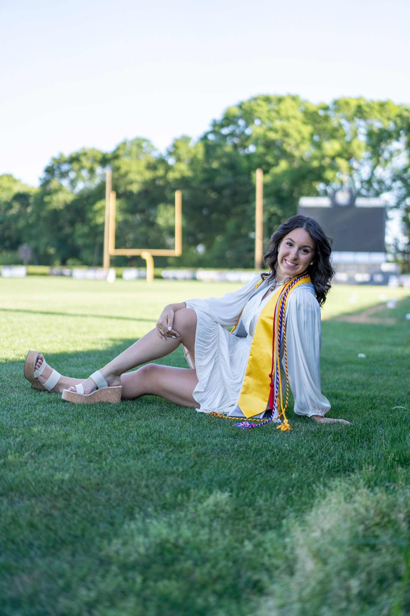 A smiling graduate sits on green grass in a white gown with gold sash, posing in front of a football field.