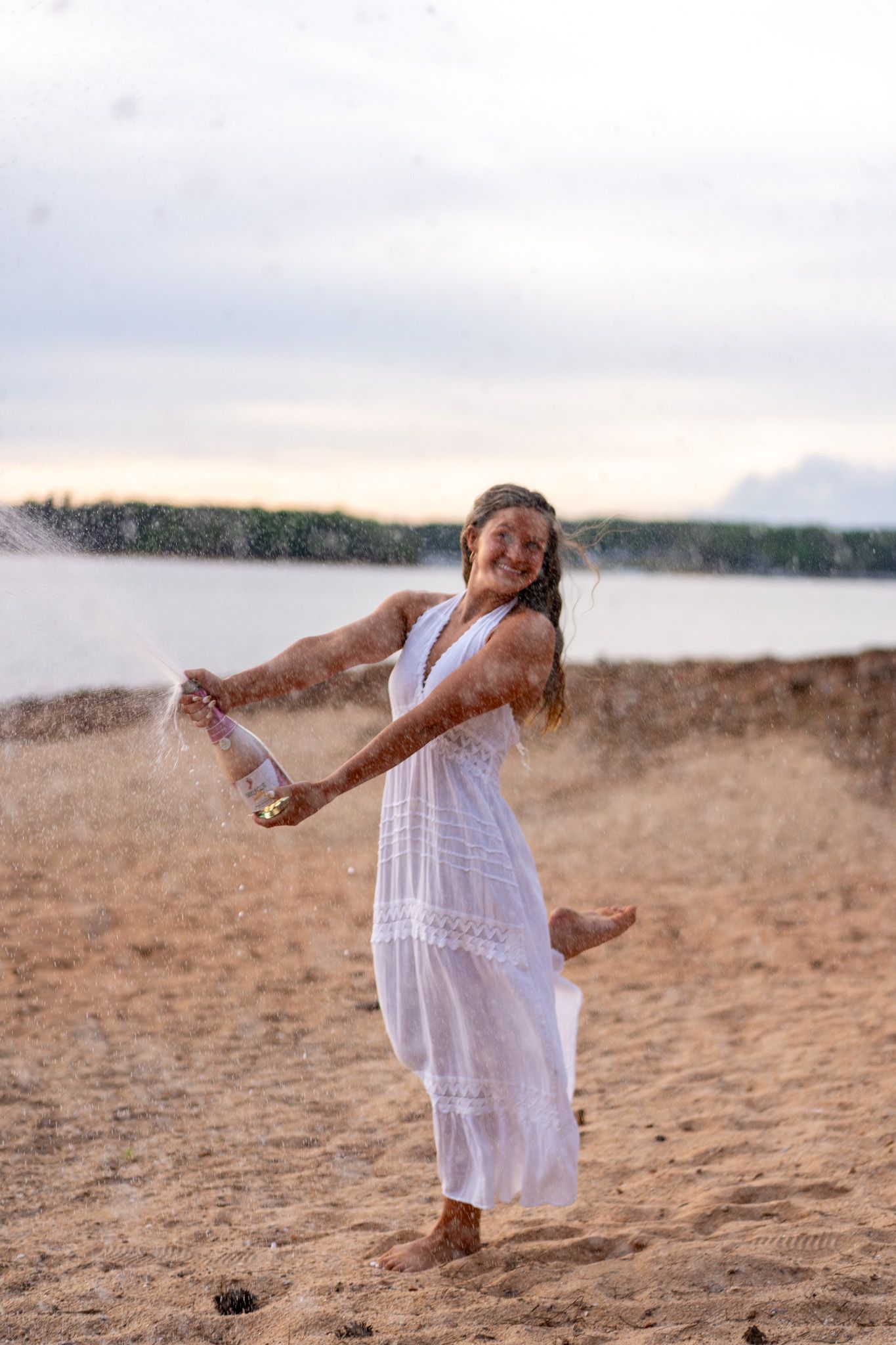 A joyful woman in a white dress celebrating on a sandy beach, spraying champagne at sunset.