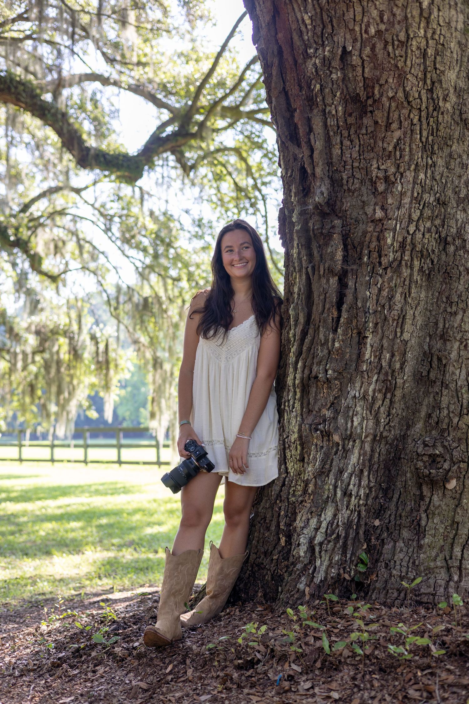 Young woman in white dress and cowboy boots holding camera, leaning against large oak tree with Spanish moss outdoors.