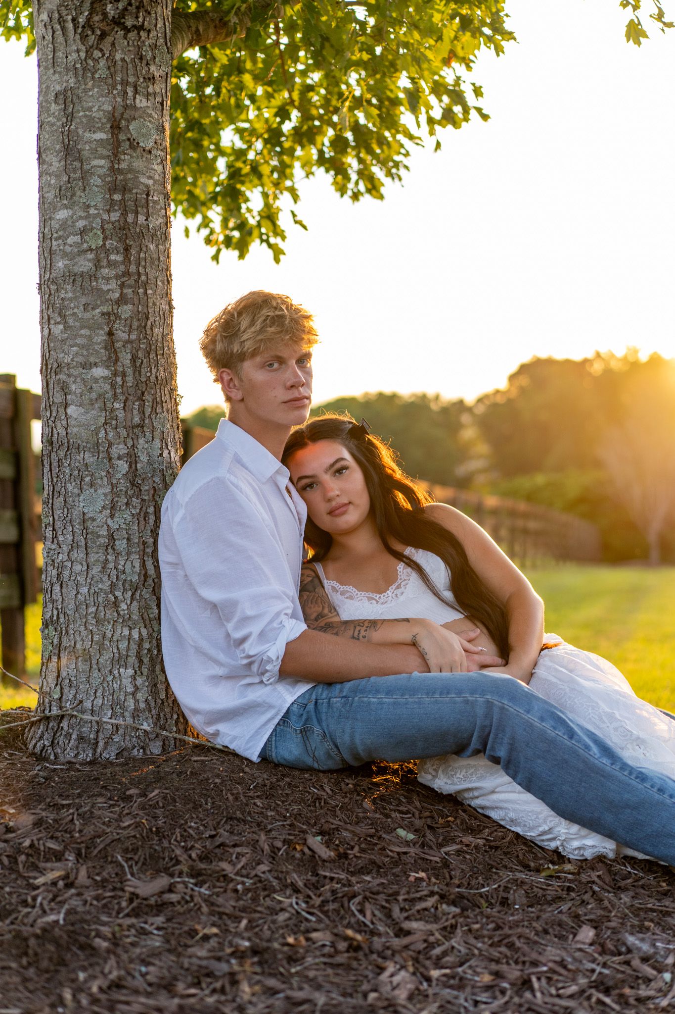 Young couple sitting against a tree at golden hour, woman in white dress leaning on man in white shirt.