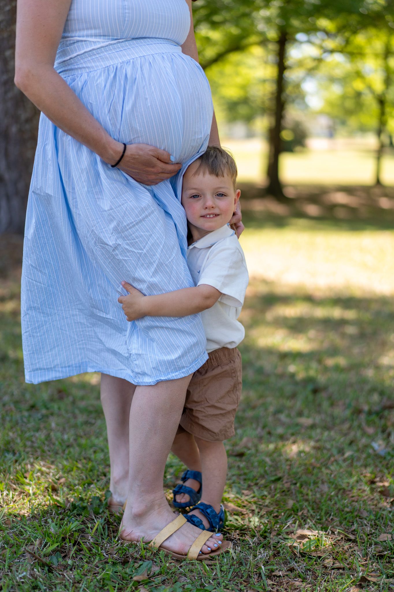 Happy toddler boy in white shirt hugs pregnant mothers baby bump outdoors in sunny green park.