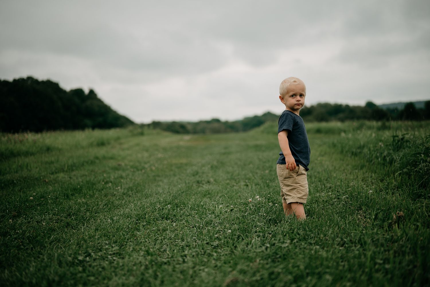 A young toddler boy in navy shirt and khaki shorts stands on a green grass path with cloudy skies.