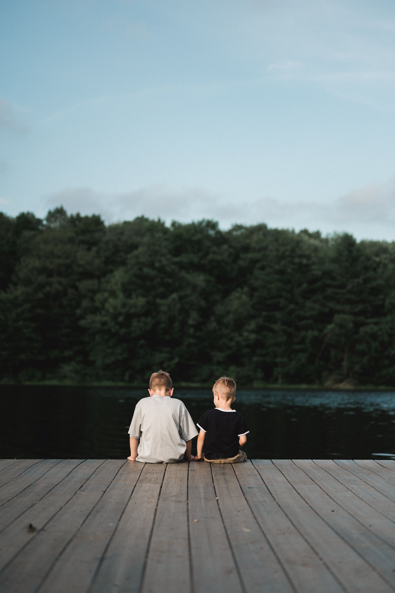Two children sitting on a wooden dock overlooking a calm lake surrounded by lush green trees.