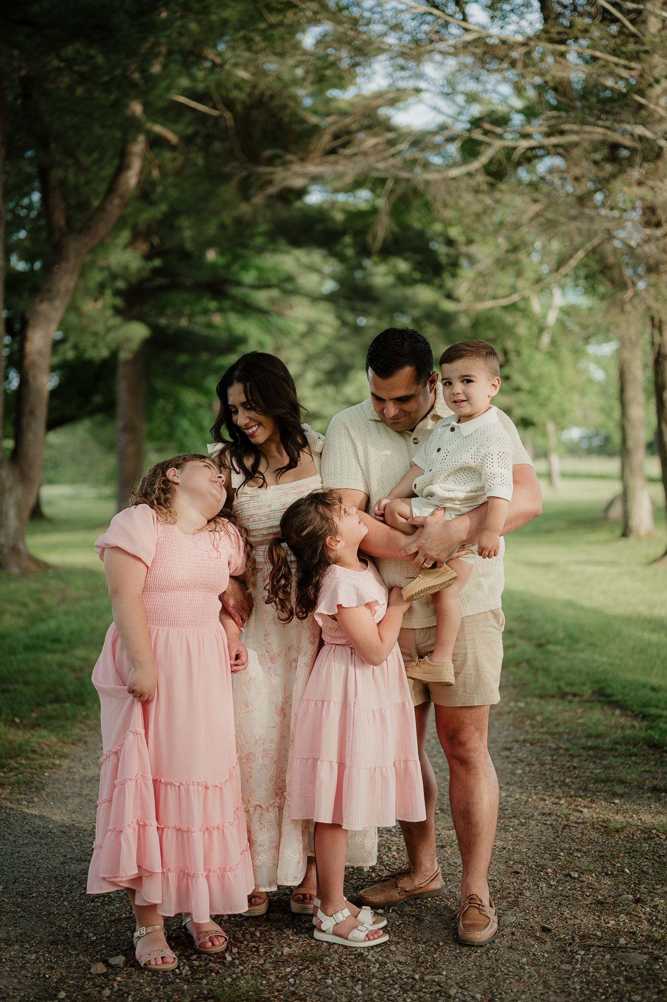 Family of five posing outdoors, parents with three young children in pink dresses and a toddler boy, surrounded by lush green trees.