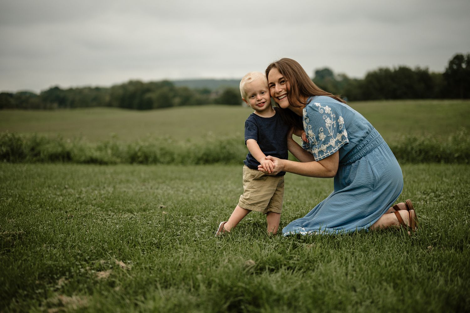 A mother kneels in a green field hugging her young blond son, both smiling outdoors on a cloudy day.