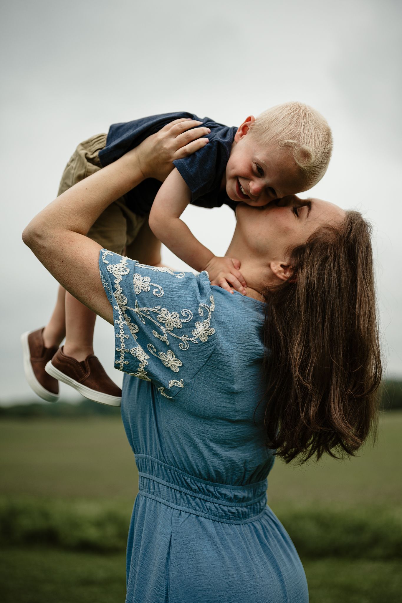 A joyful mother in a blue dress lifts her laughing toddler son outdoors in a green field.