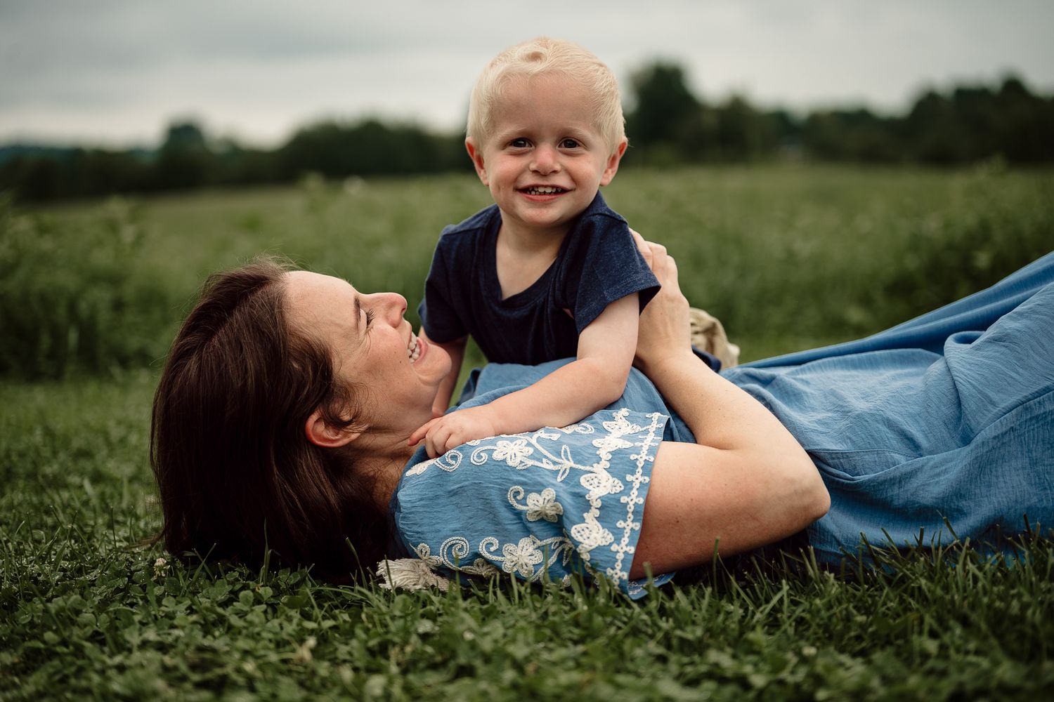 A mother lies on green grass laughing as her toddler boy smiles and plays on top of her outdoors.