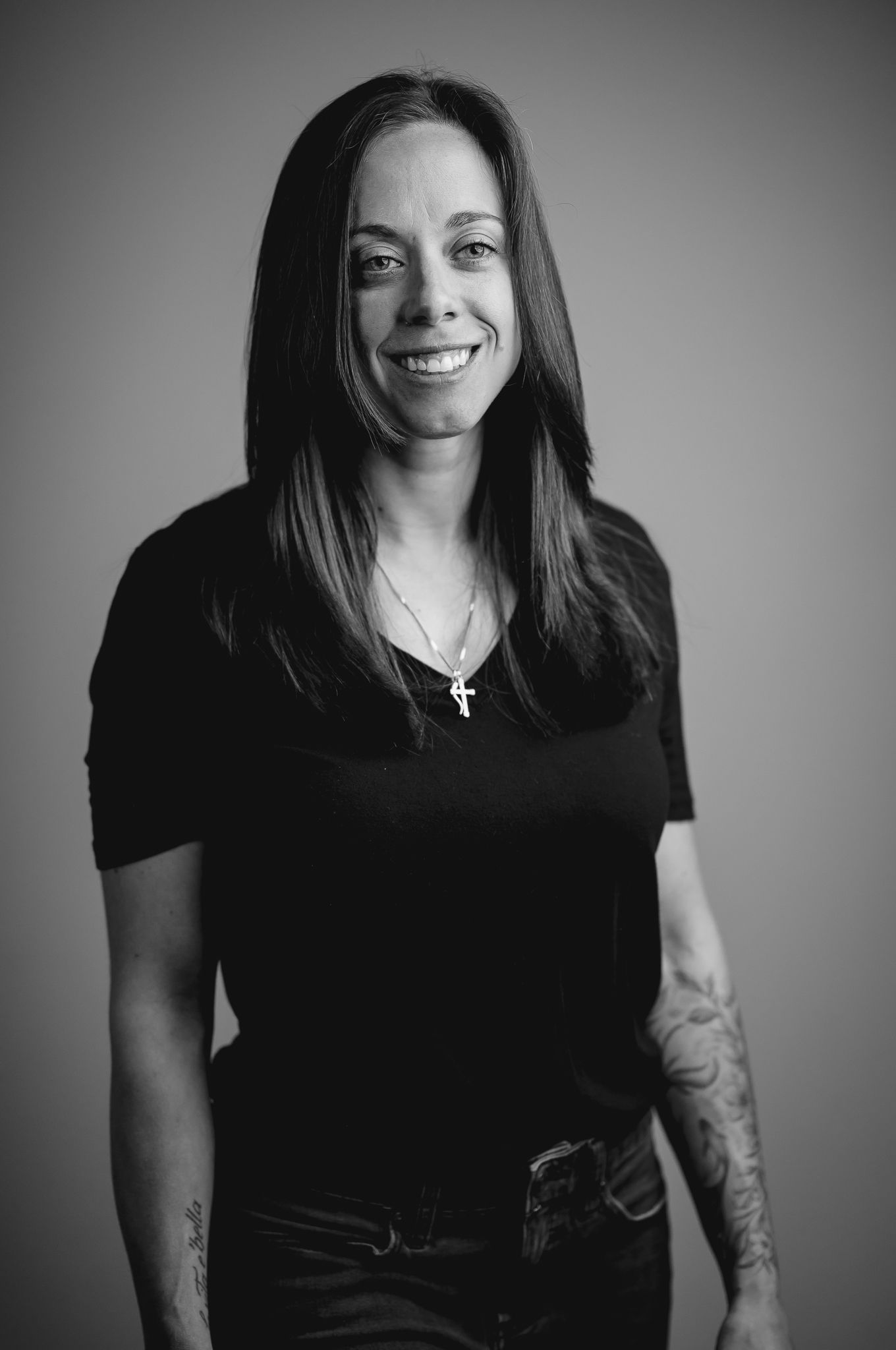 Smiling woman with long dark hair, cross necklace, black t-shirt, and tattoos poses for a black and white portrait.