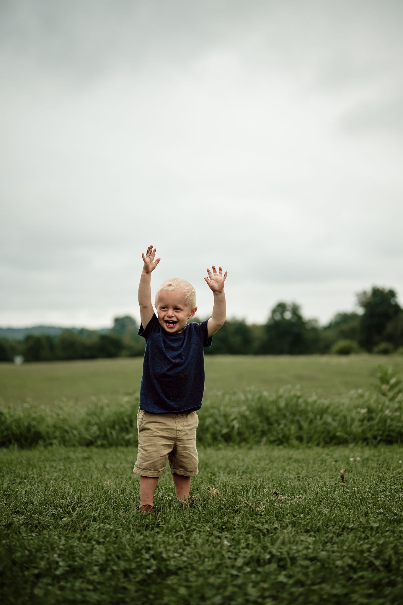 A joyful toddler boy raises his hands in a green field, wearing a navy shirt and khaki shorts on a cloudy day.