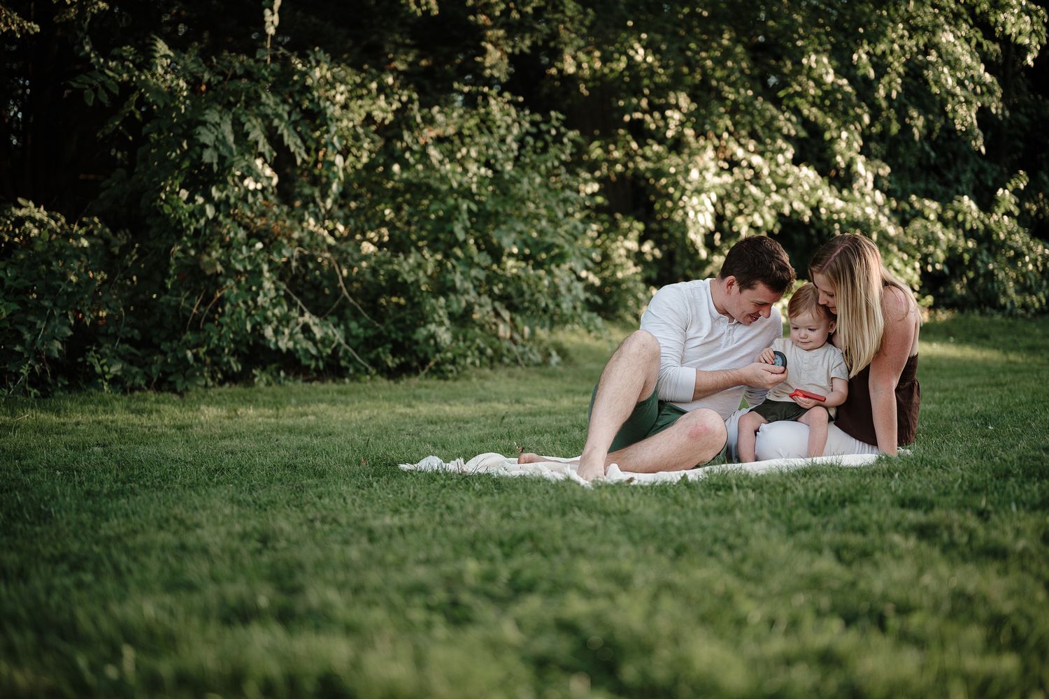 A young family sits together on a blanket in a lush green park, parents lovingly focused on their toddler.