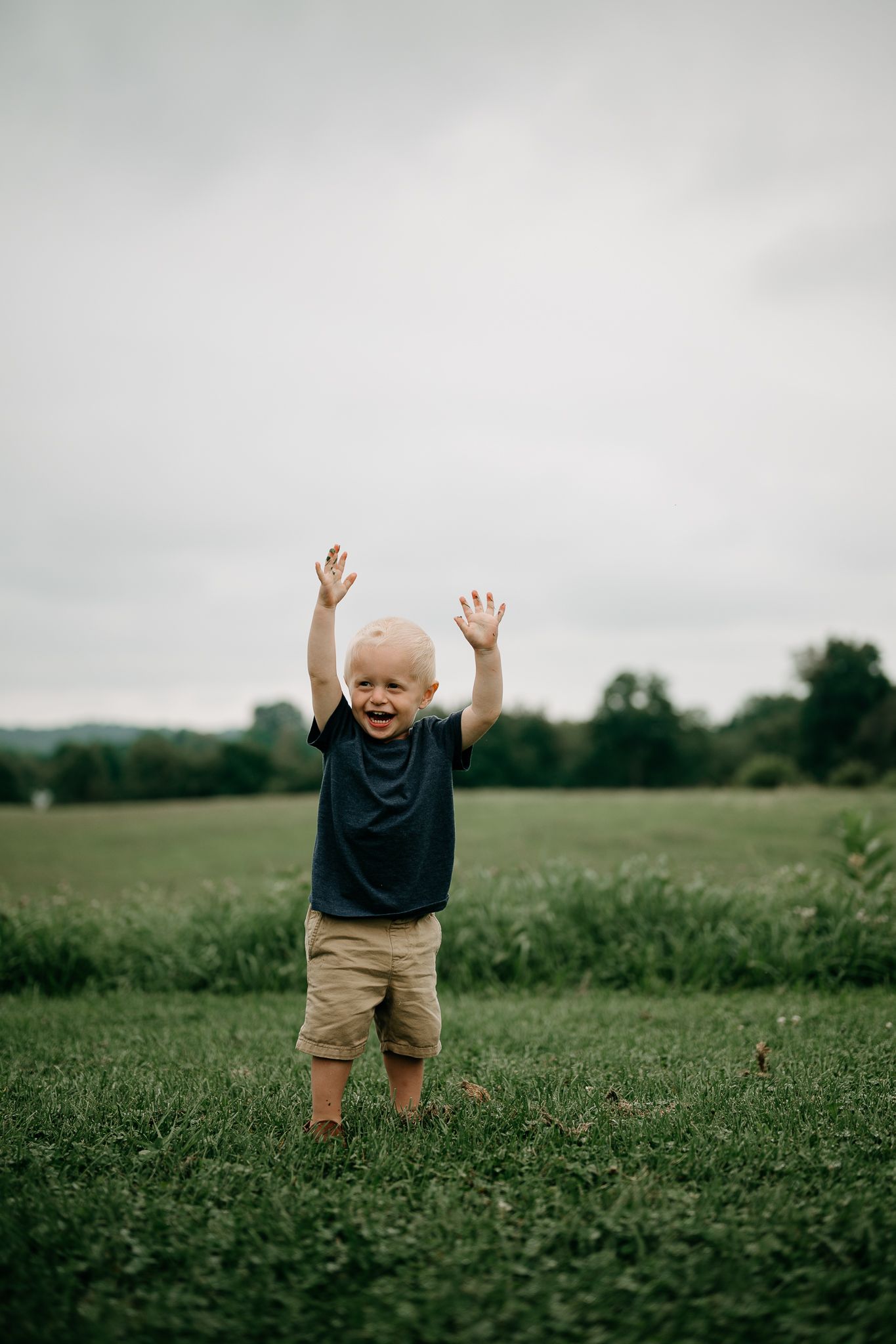 A joyful toddler in a navy shirt and tan shorts stands in a green field with arms raised, smiling outdoors.