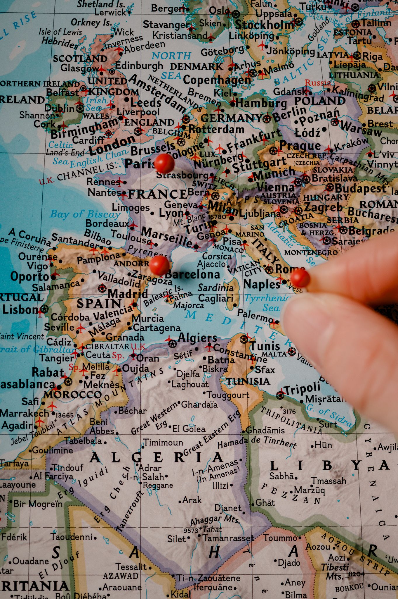 A hand points to red pins marking locations across a colorful map of Europe and North Africa.