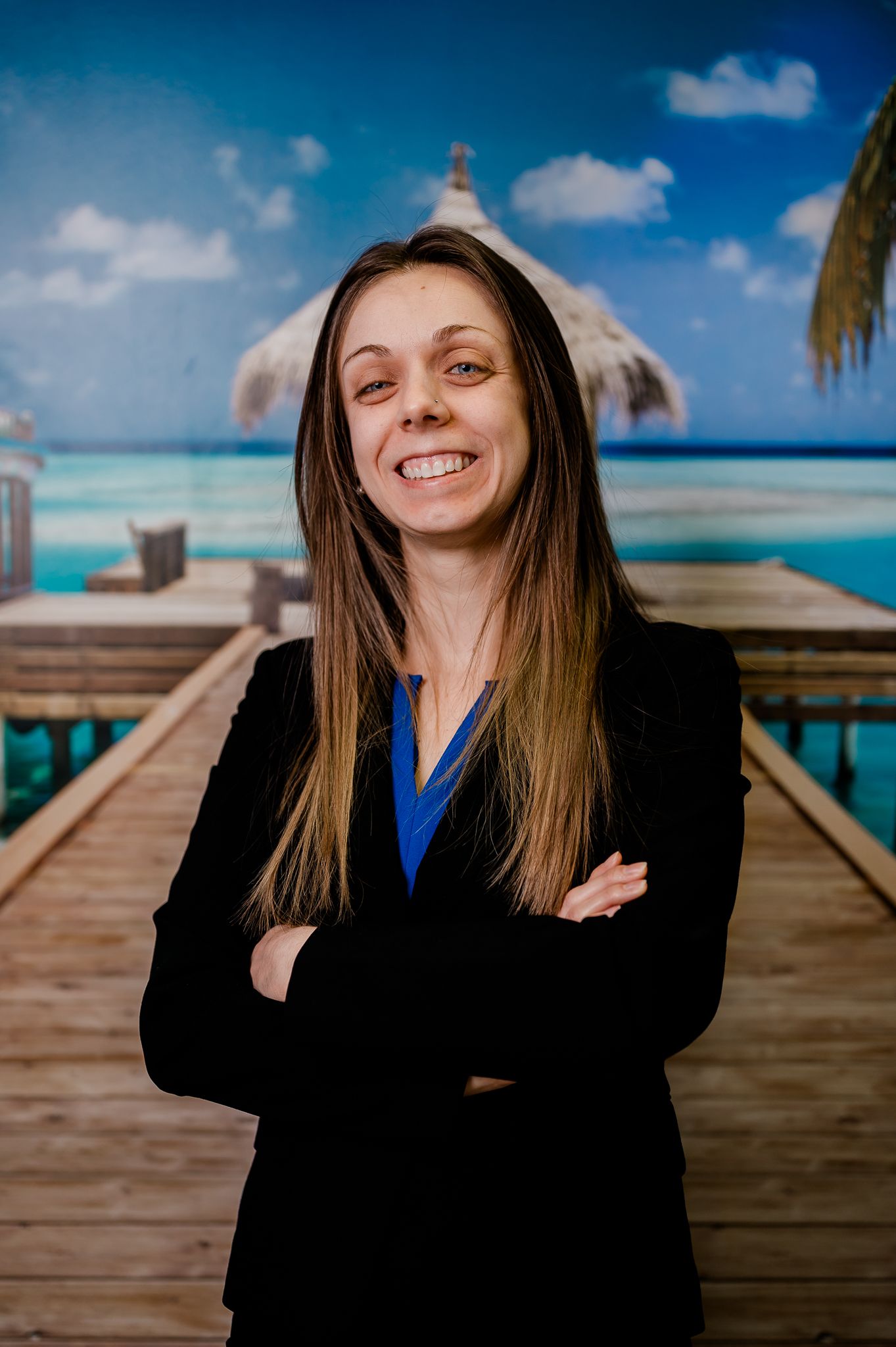 Smiling woman in black blazer stands with arms crossed before tropical beach backdrop.