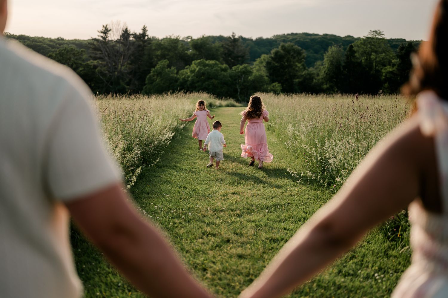 Parents holding hands watching three children run through lush green meadow path.