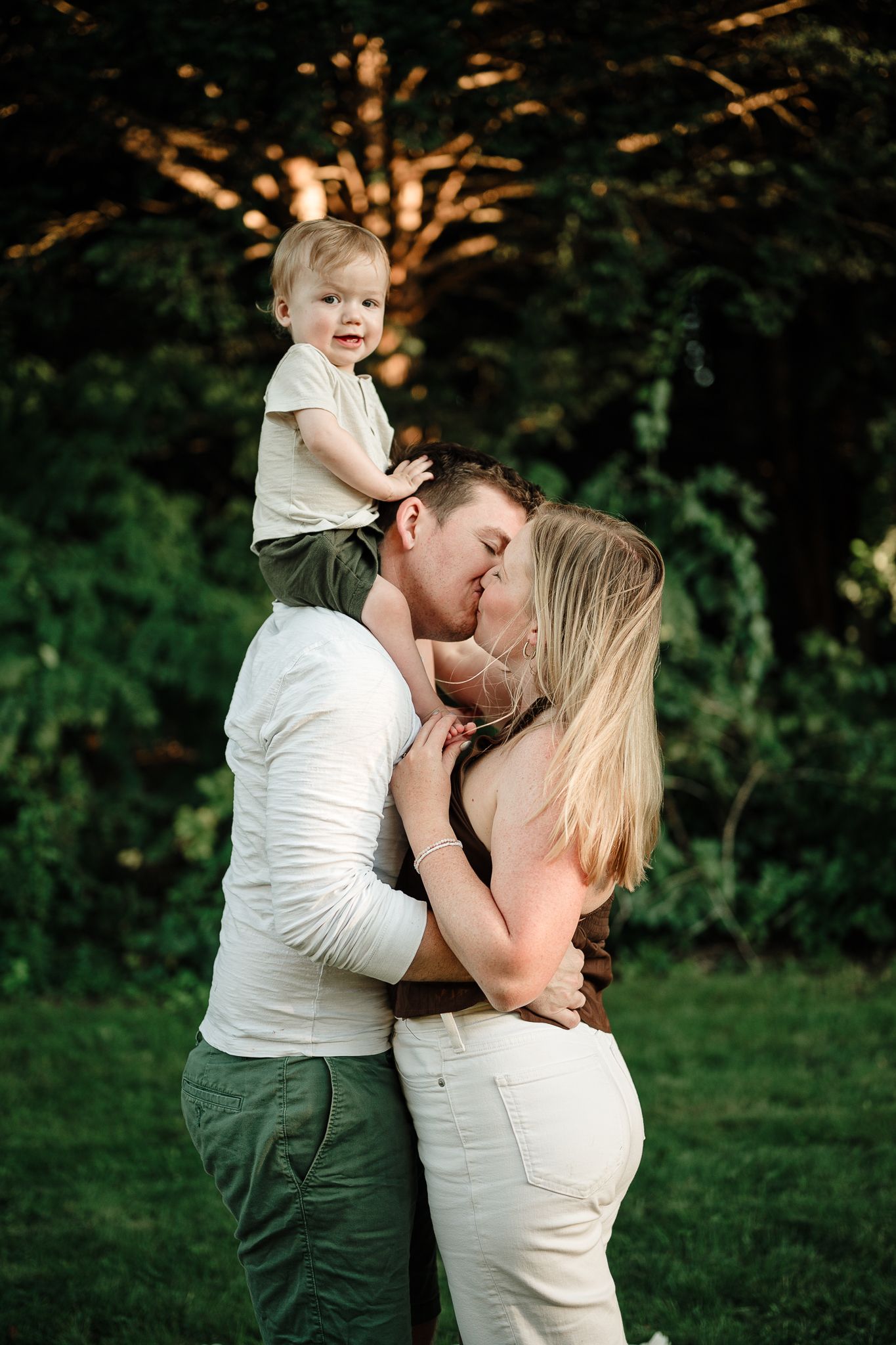 A couple shares a kiss outdoors while their toddler sits on dad's shoulders, smiling at the camera.