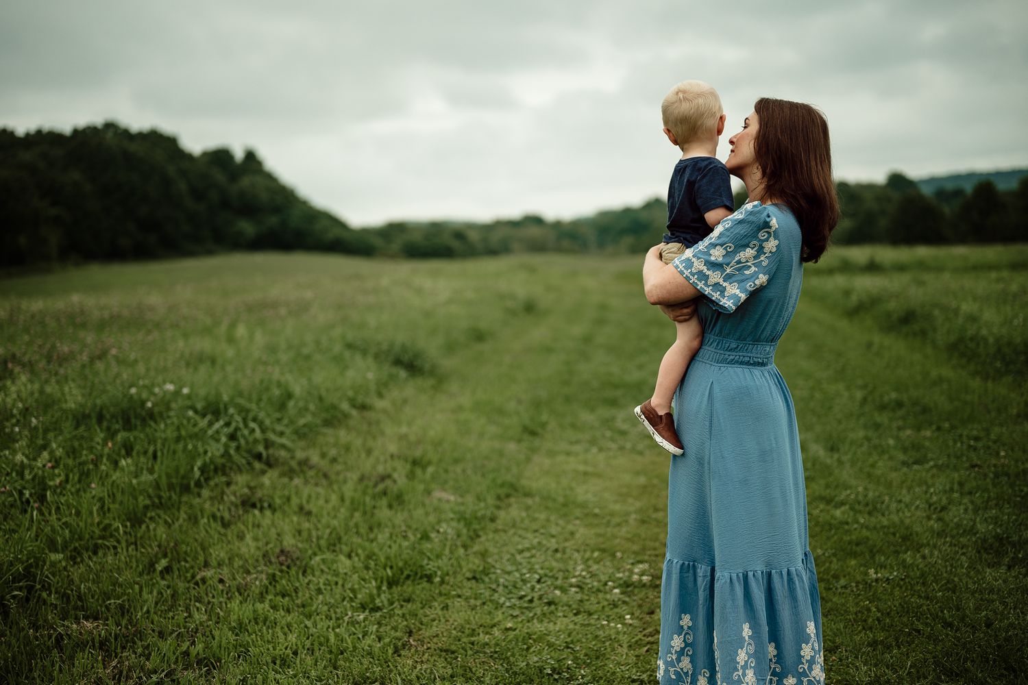 A mother in a blue floral dress holds her toddler in a lush green field under an overcast sky.