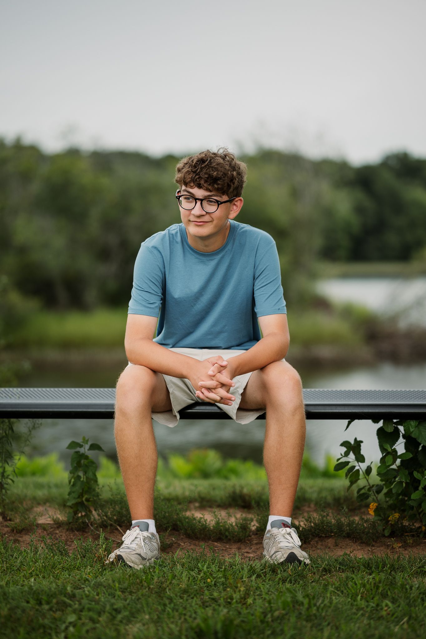 Teen boy with curly hair and glasses sits on a bench outdoors, wearing a blue t-shirt and khaki shorts.