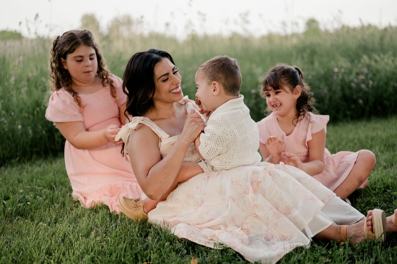 A mother in a floral dress sits in grass with her three young children at sunset.