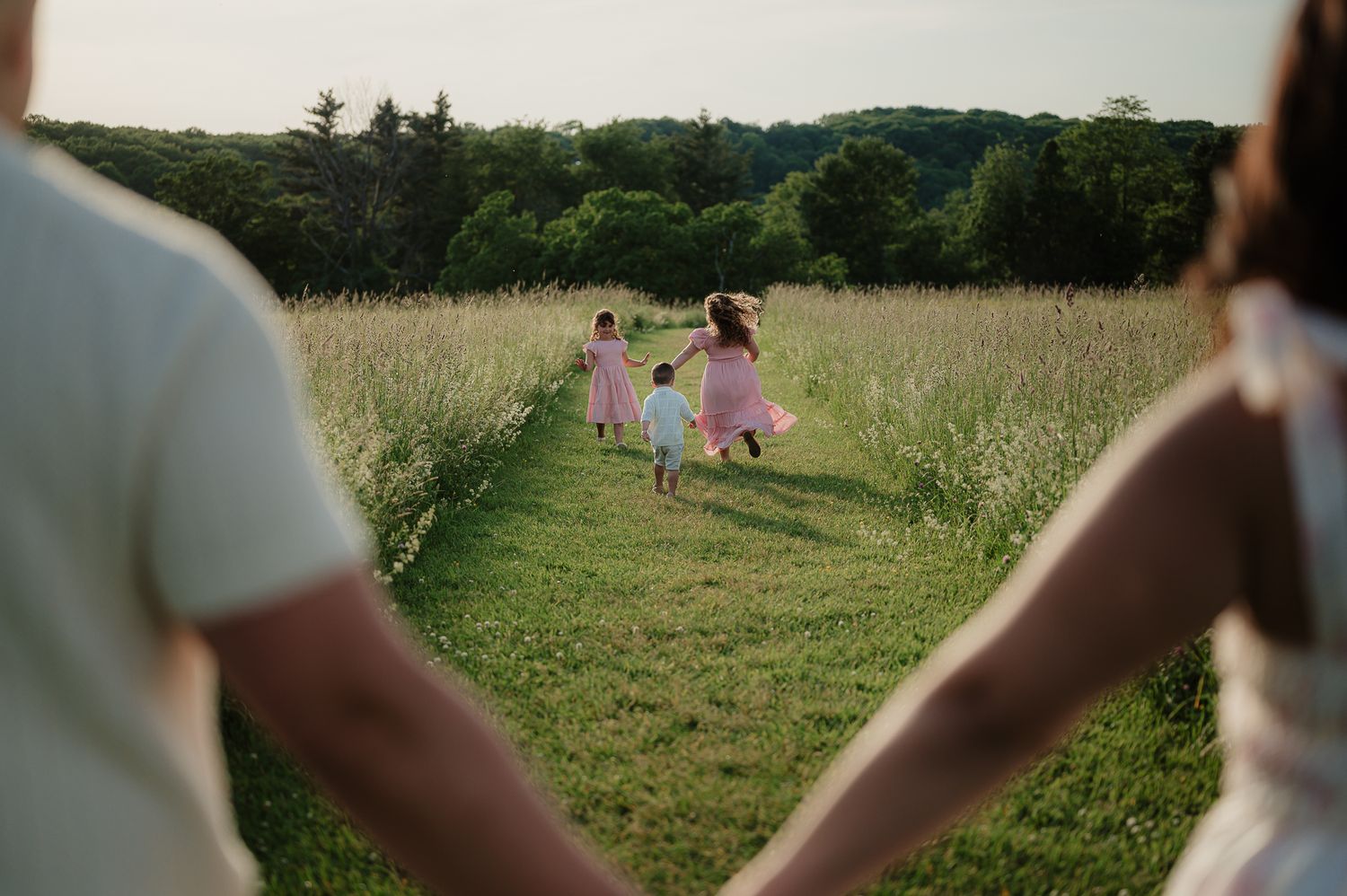 Children in pink dresses run through a lush green meadow while parents hold hands in the foreground.