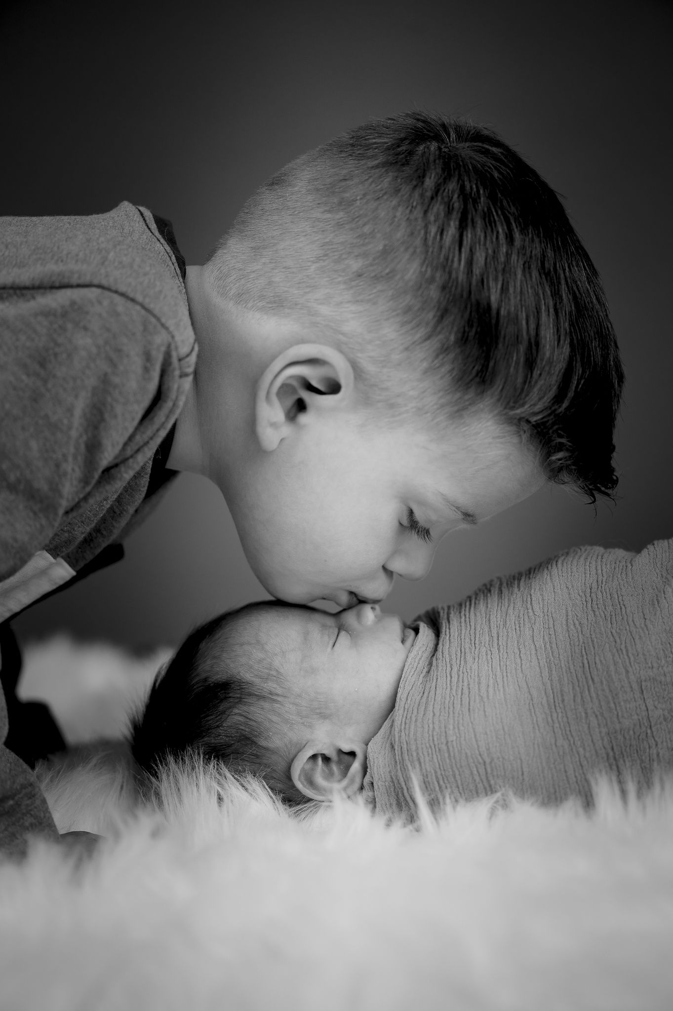 Tender black and white photo of young boy gently kissing sleeping newborn baby sibling.