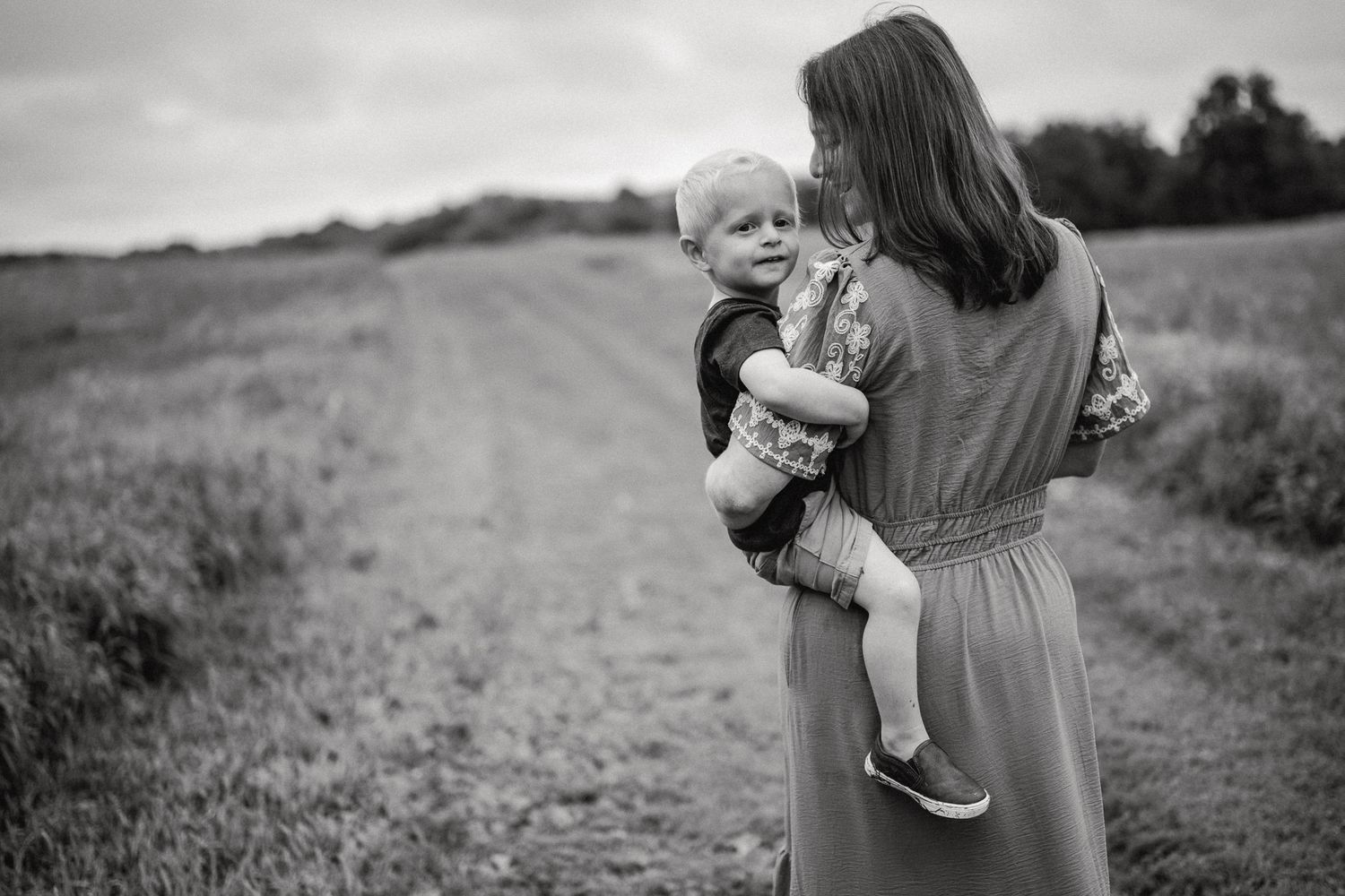 Black and white photo of a mother holding her toddler on a rural dirt path surrounded by fields.