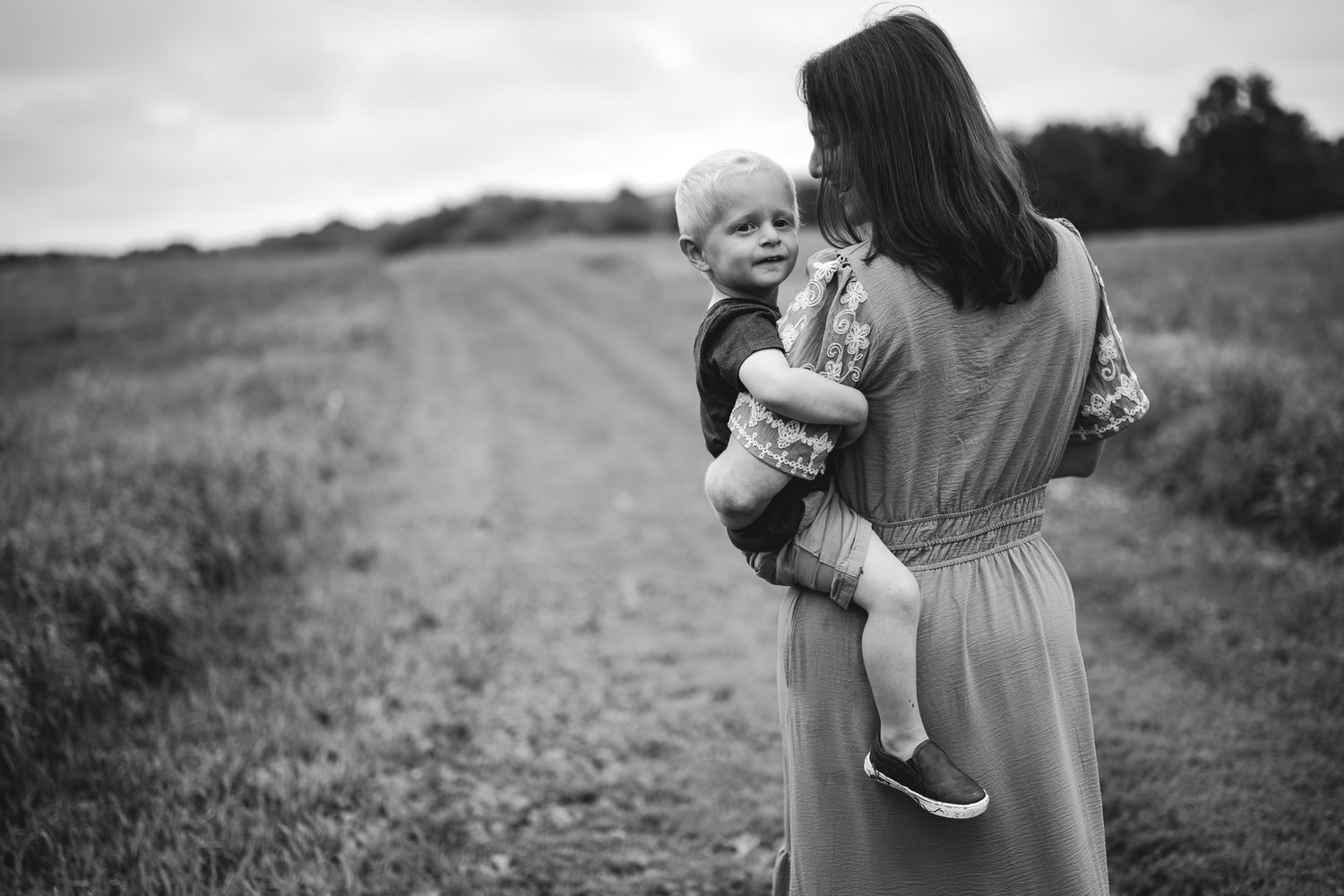 Black and white photo of a mother holding her toddler in a field, both facing away from the camera.