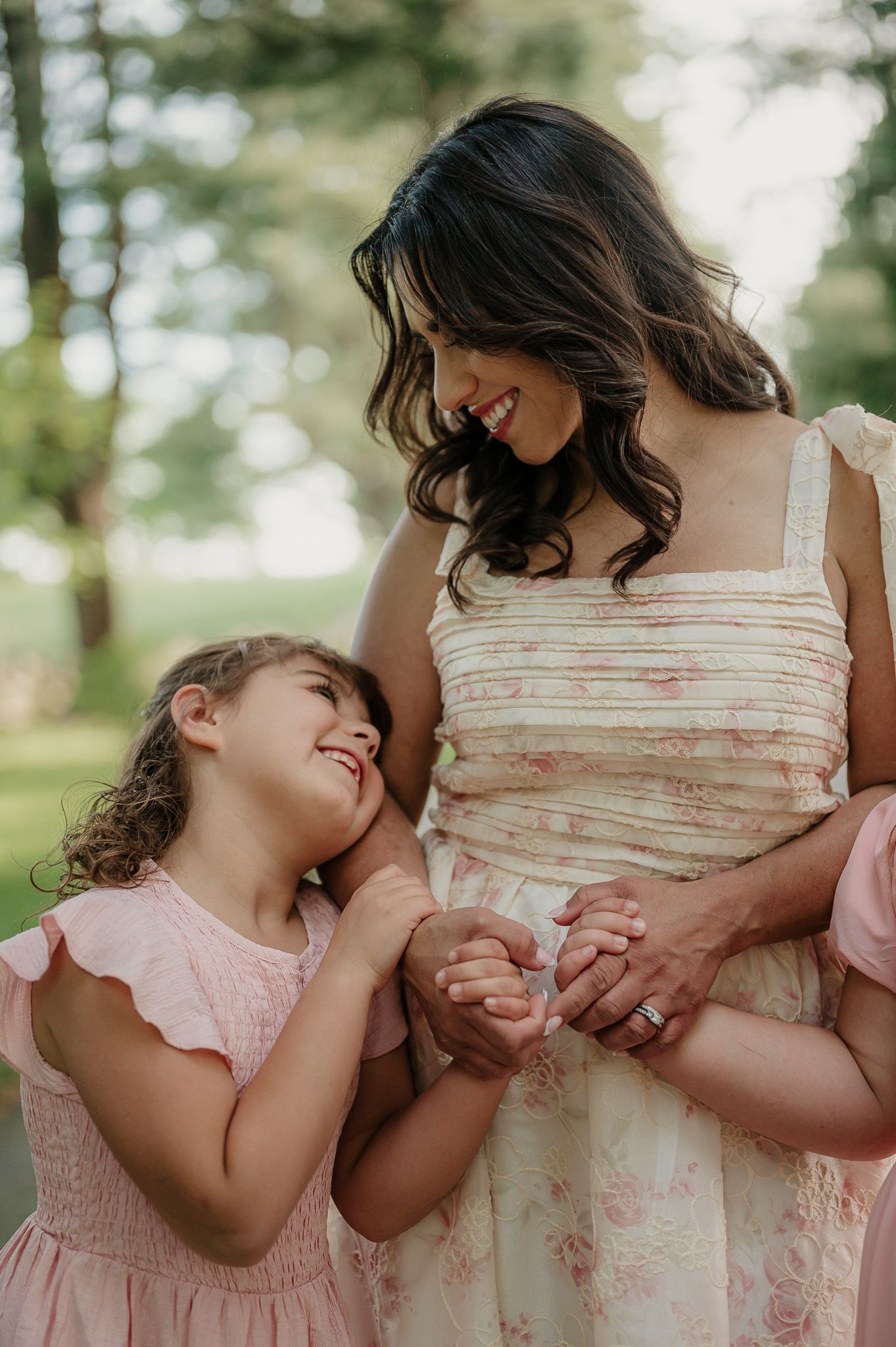 A mother and young daughter in pink floral dresses share a joyful moment outdoors, smiling and holding hands.