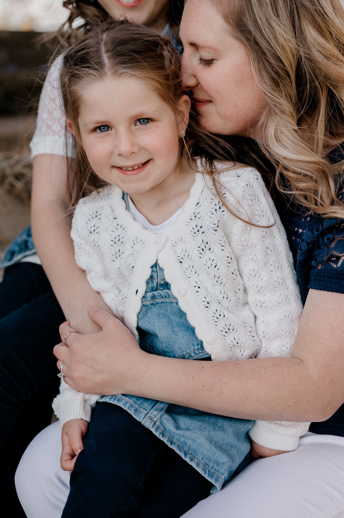 Mother kissing young blonde girl on cheek while hugging her warmly outdoors.