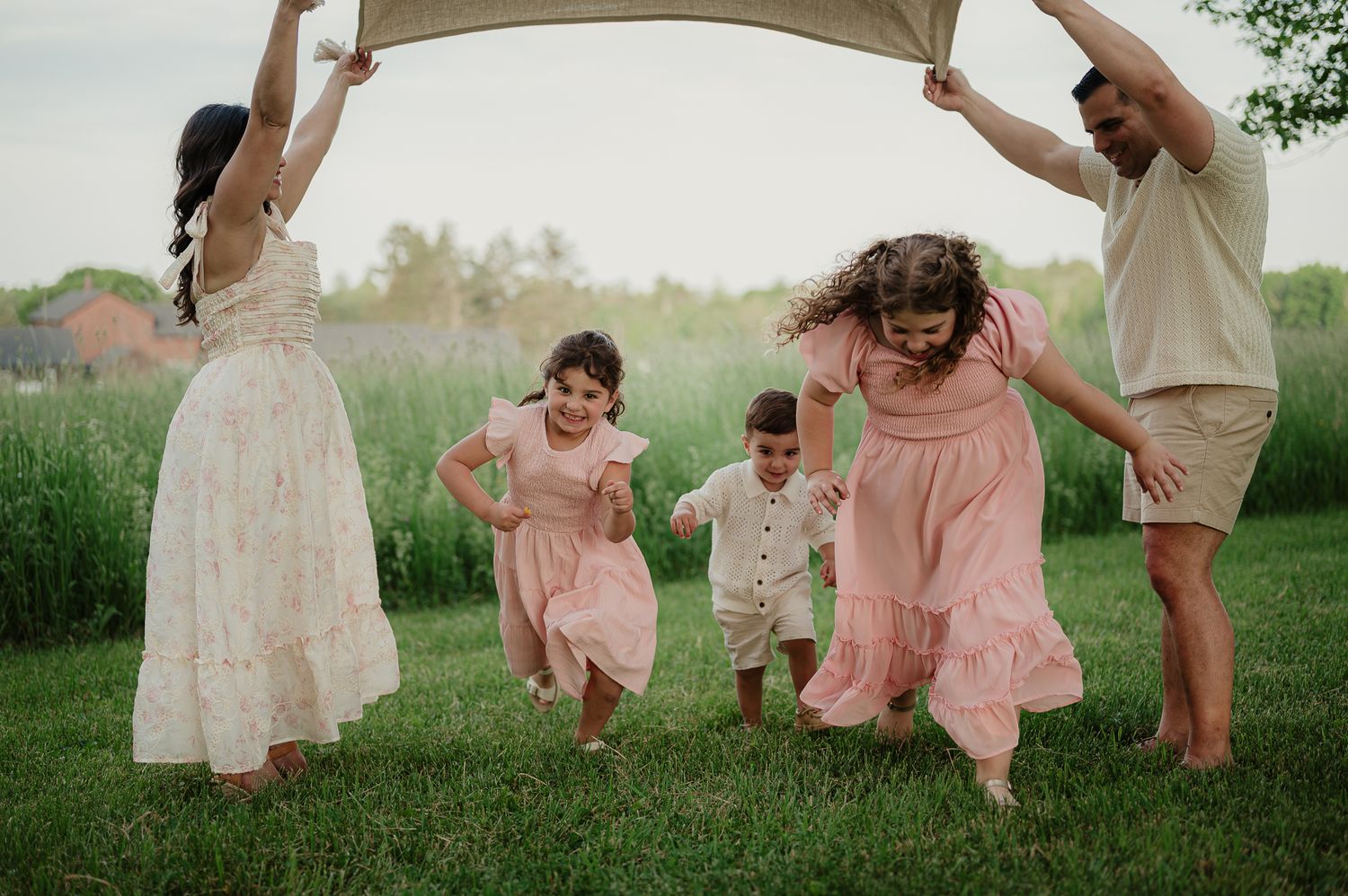 Family of five playing under a blanket on green grass, parents and three children in pink and white outfits.